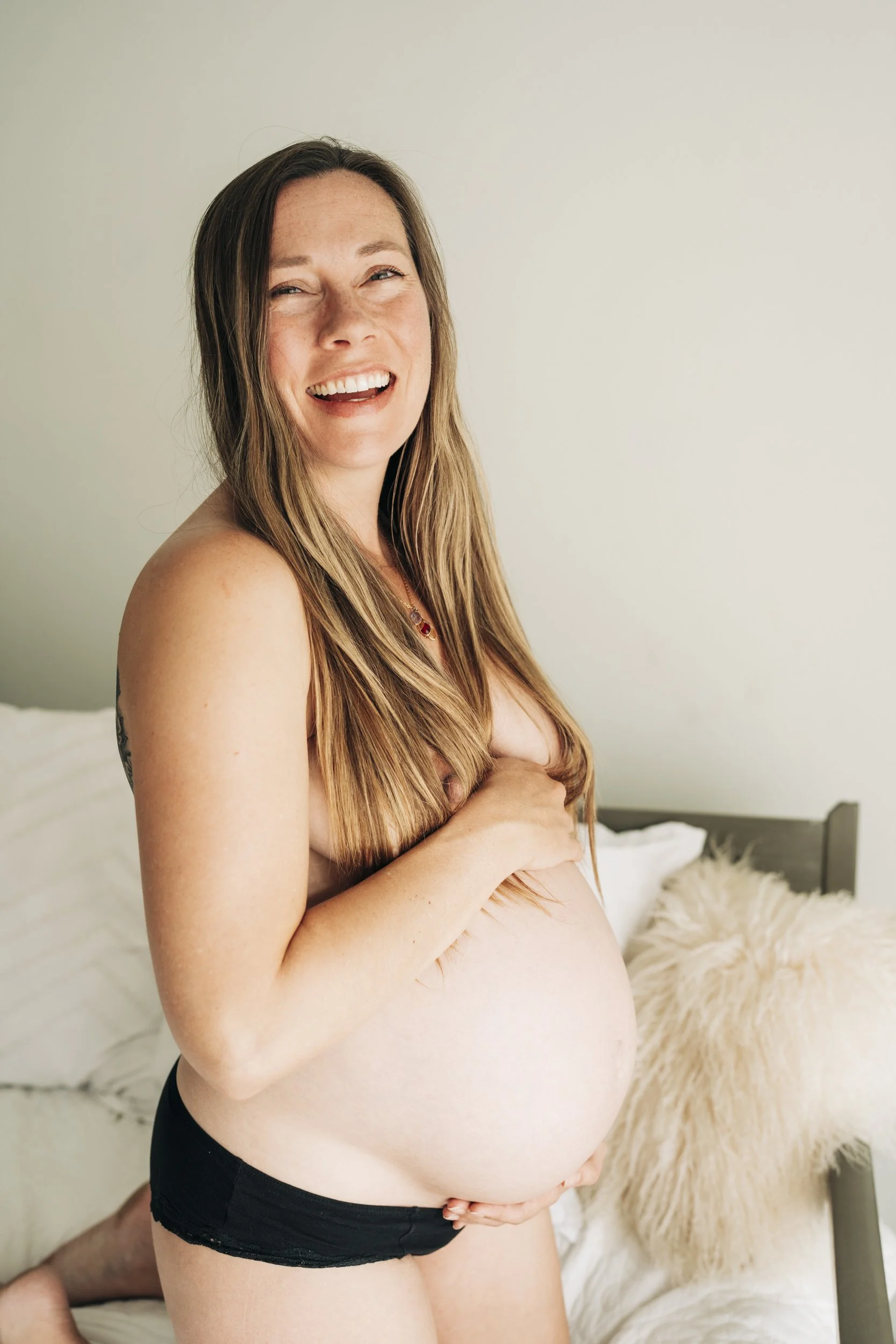 A pregnant woman smiling and covering her breasts with her hand, sitting on a bed with white bedding and a fluffy cream-colored pillow near a neutral wall.