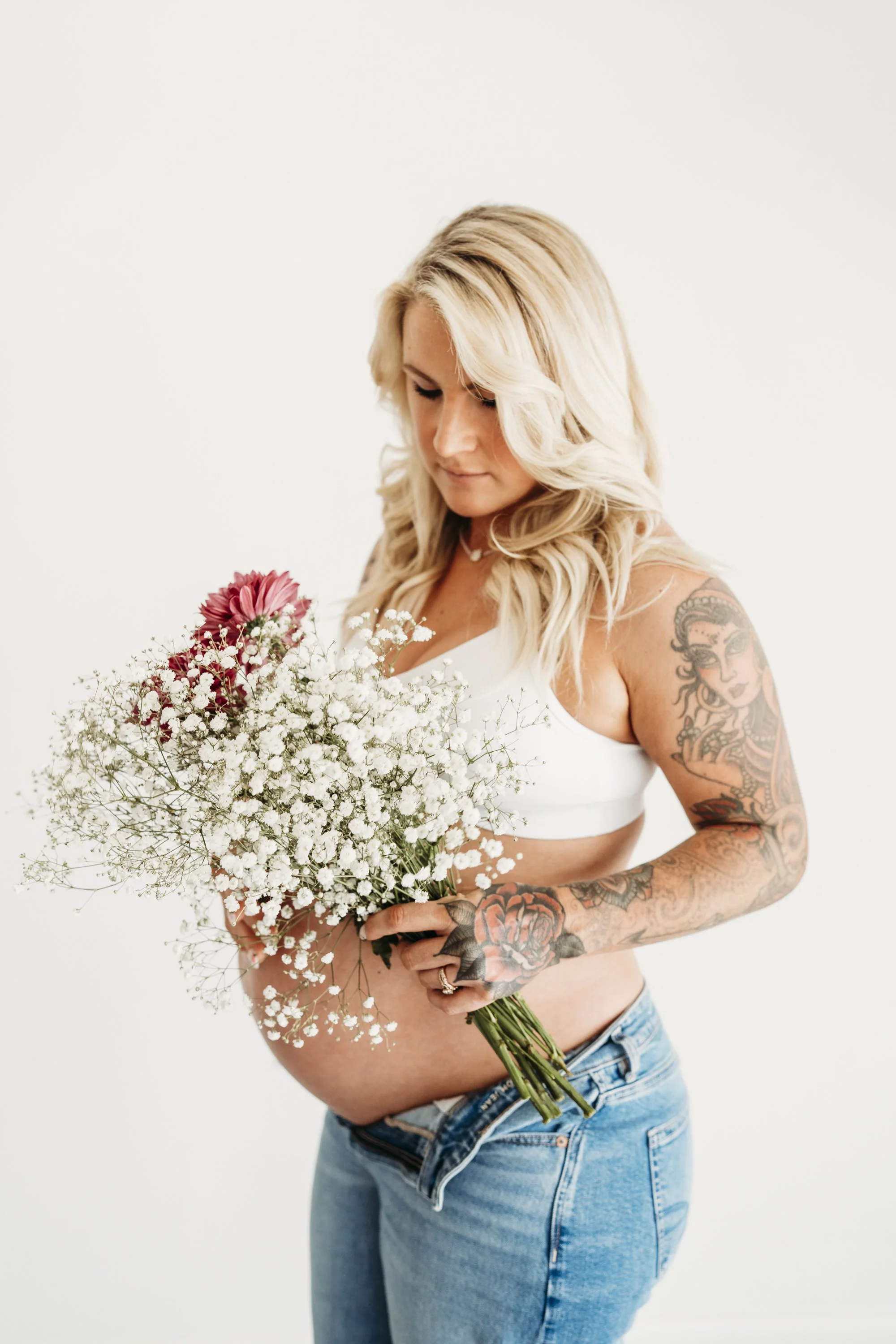 Pregnant woman with blonde hair, tattoos on arms and chest, holding a bouquet of flowers, standing against a plain white background.