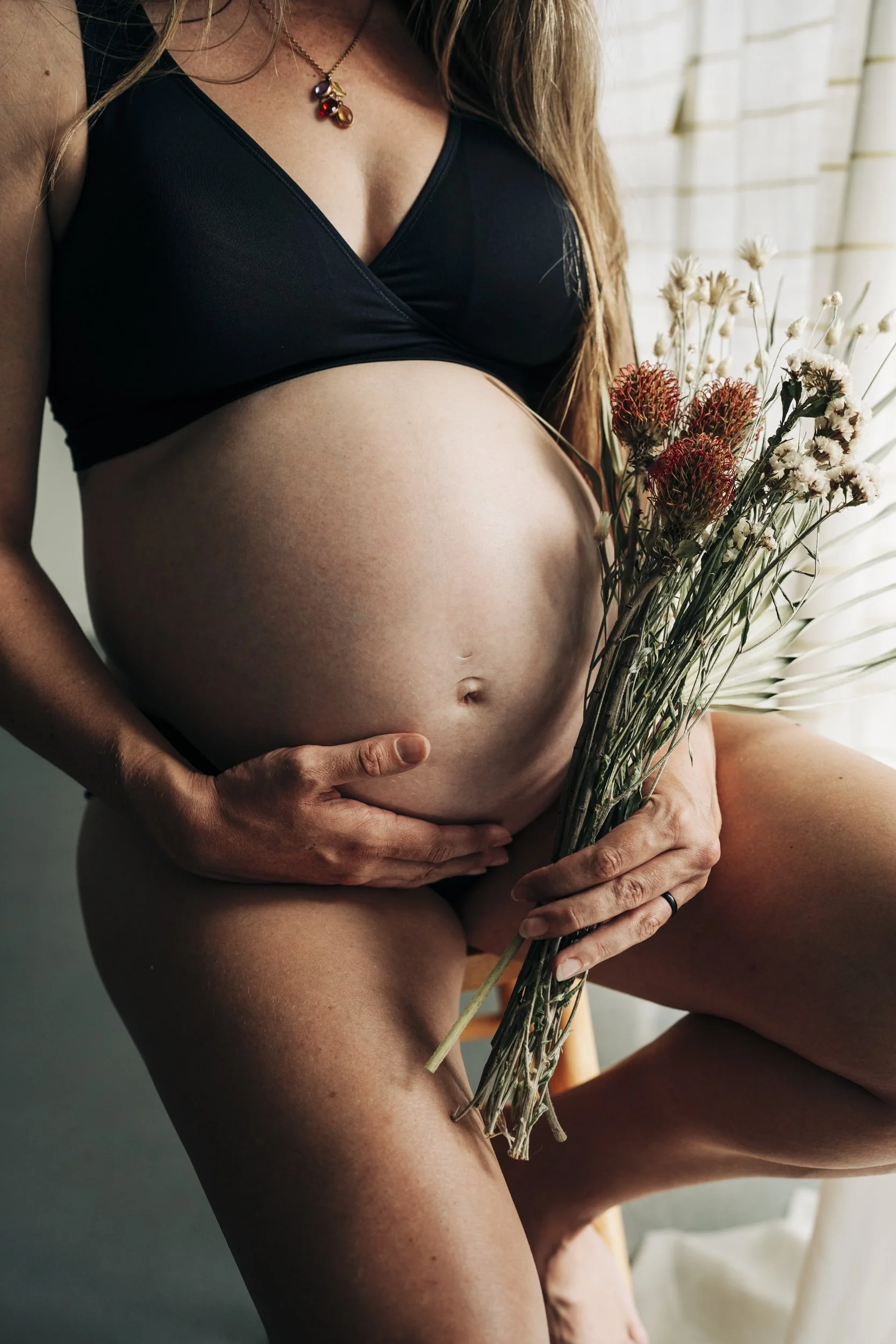 A pregnant woman holding a bouquet of dried flowers, wearing a black bra, with her hand on her belly.