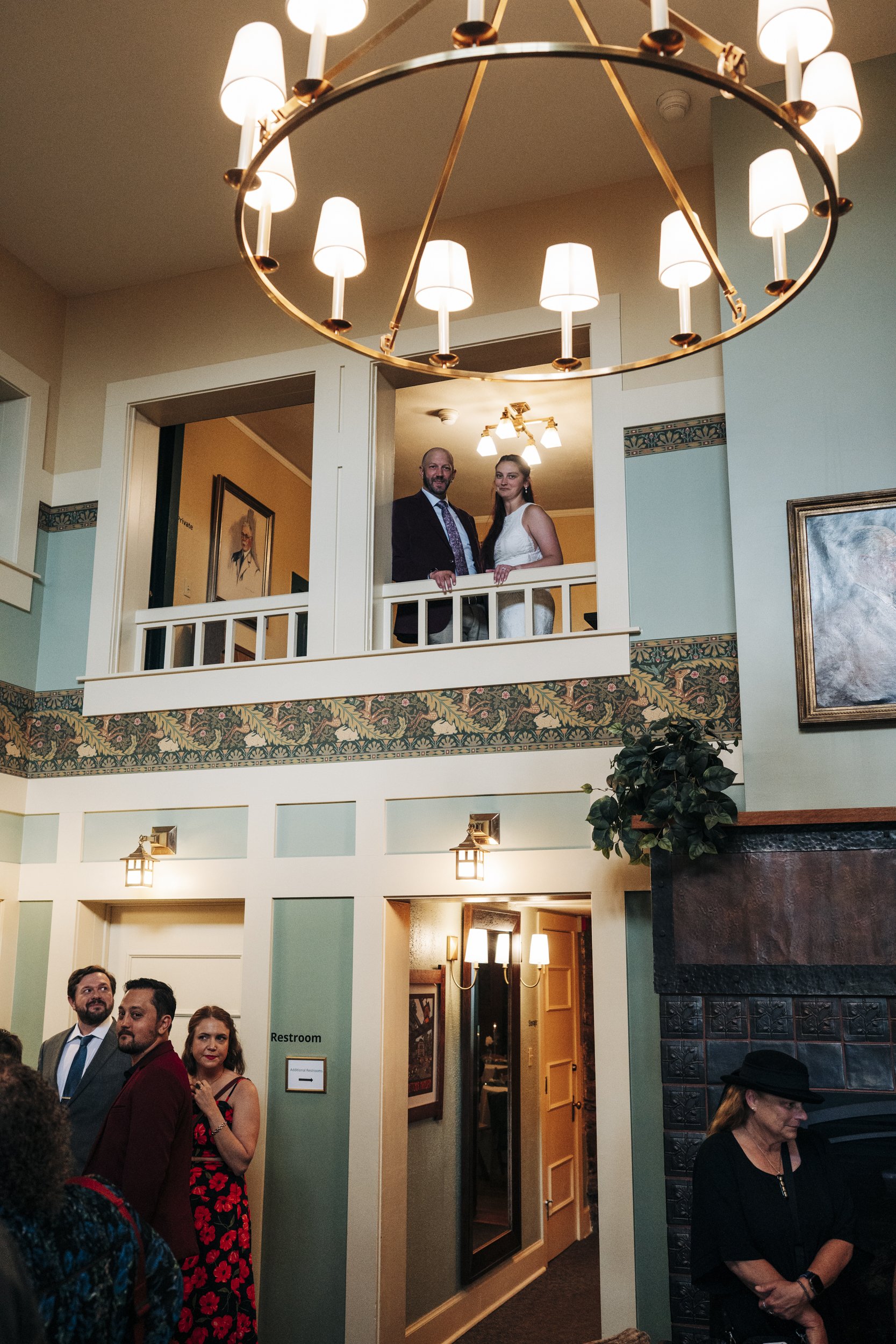 People gathered in a building with two levels, a chandelier hanging from the ceiling, and a mix of interior decor including lamps, framed pictures, and a sign indicating the restroom. Two individuals stand on the upper level balcony, looking down, wh