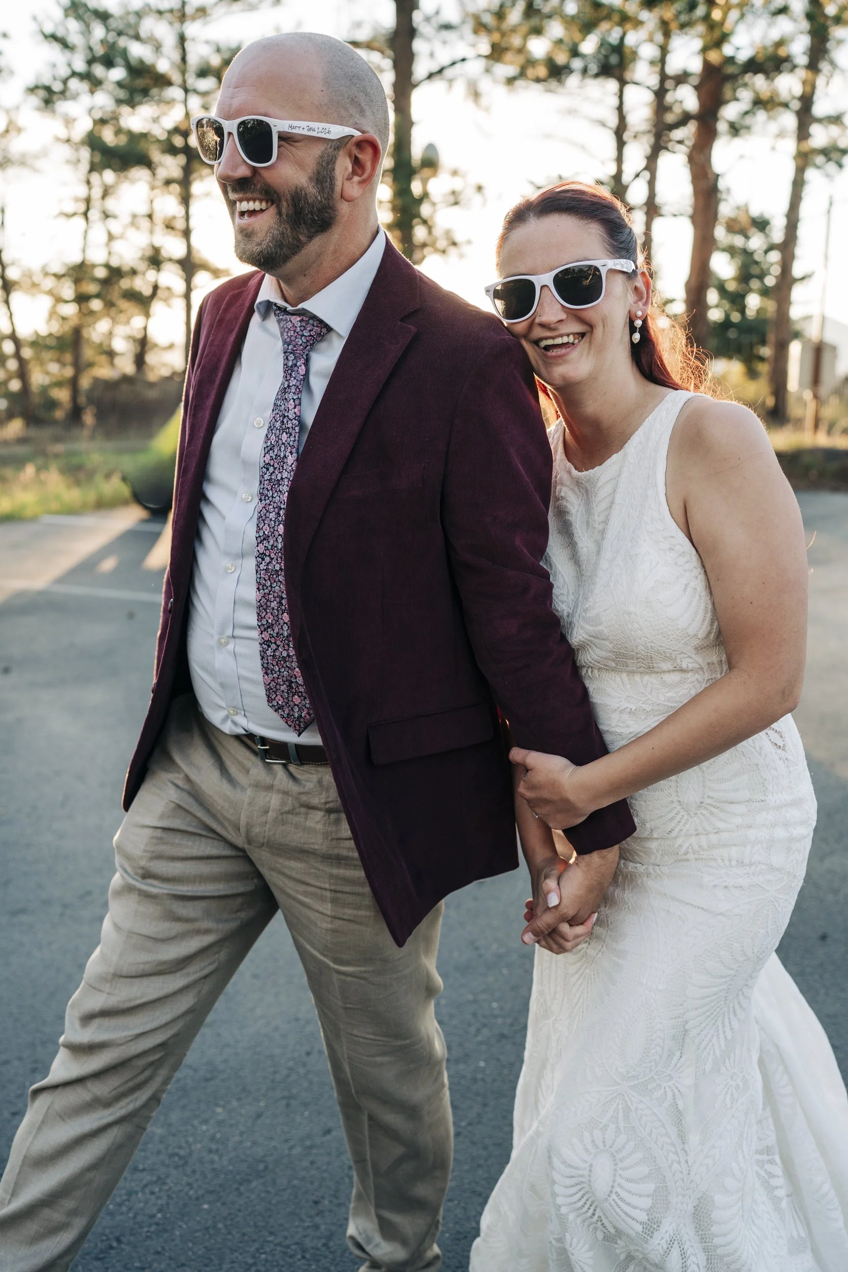 A smiling couple holding hands and walking outdoors, dressed in stylish attire, with trees and sunlight in the background.