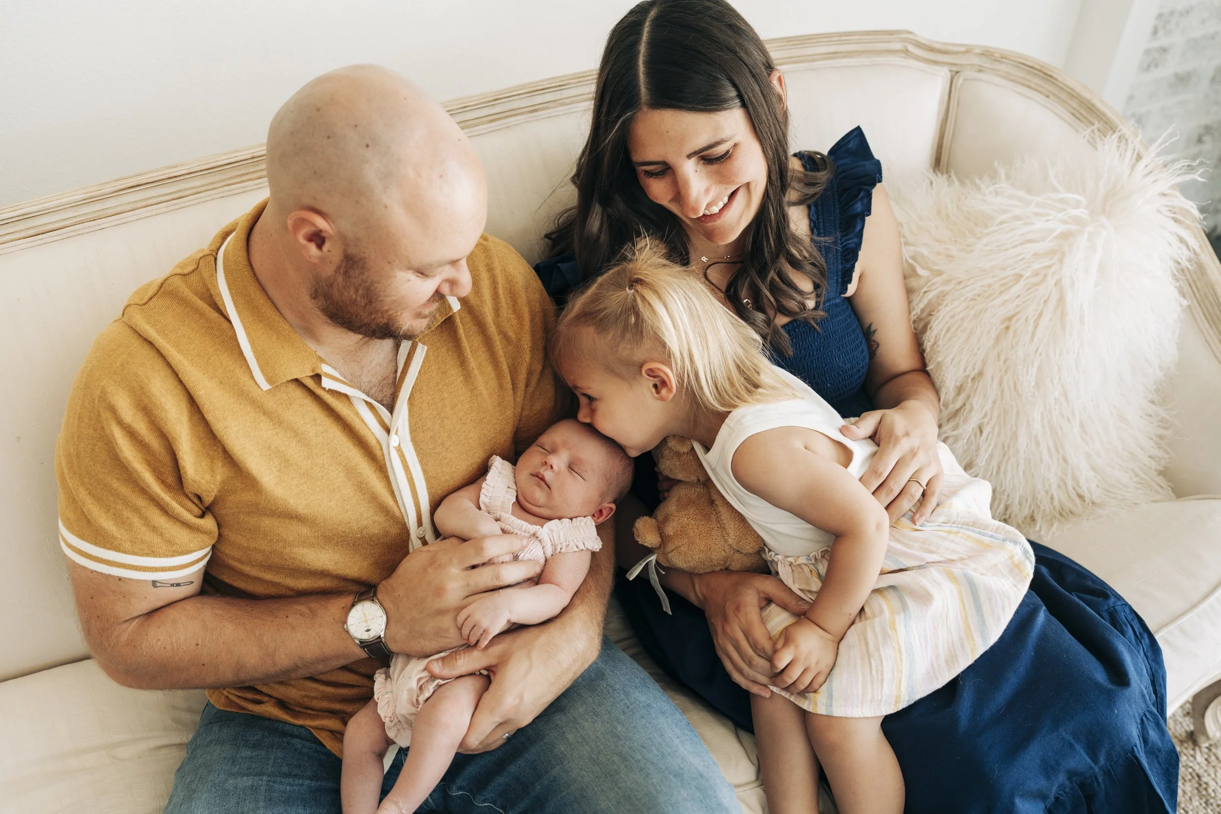 A family of four sitting on a cream-colored sofa, with the father holding a newborn baby, the mother smiling, and a young girl kissing the baby on the forehead.