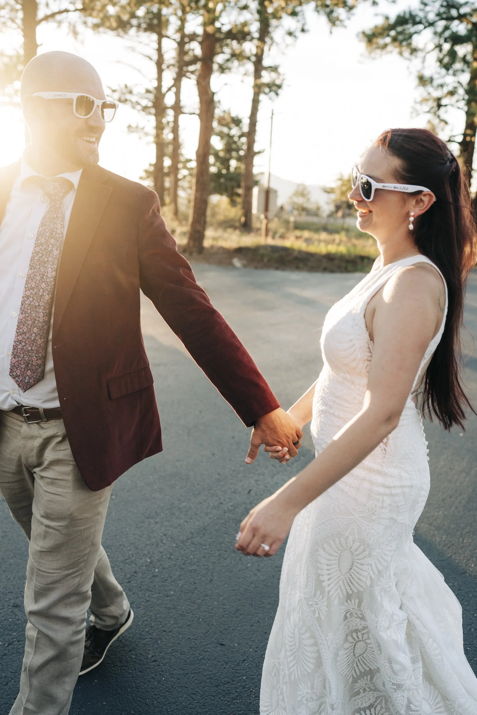 A man and woman holding hands outdoors in a sunny, wooded area. The woman is wearing a white lace dress and the man is dressed in a burgundy jacket with a patterned tie. They are smiling and wearing sunglasses.