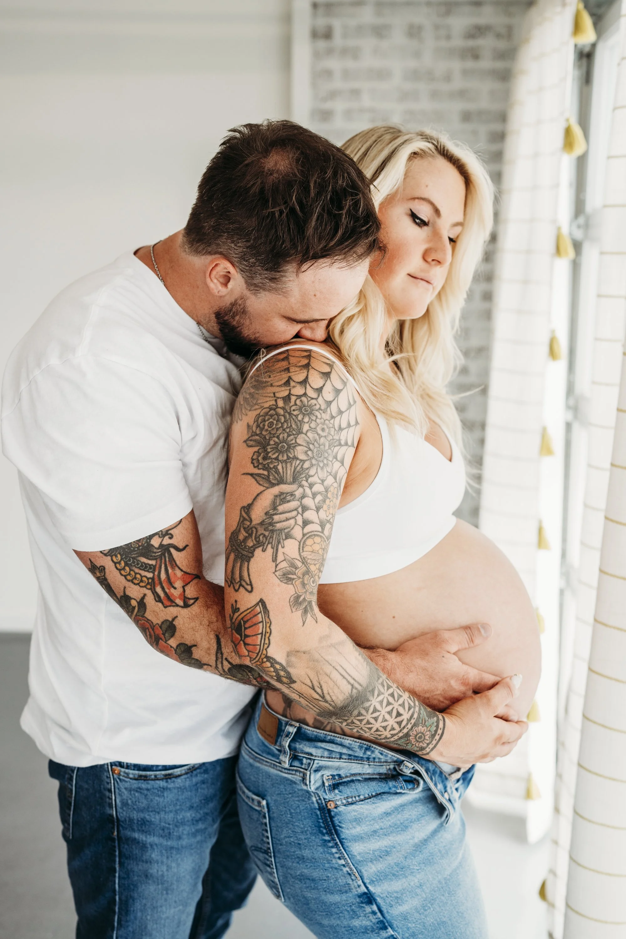 Pregnant woman with blonde hair and tattoos on her arm looking out window, with man embracing her from behind.