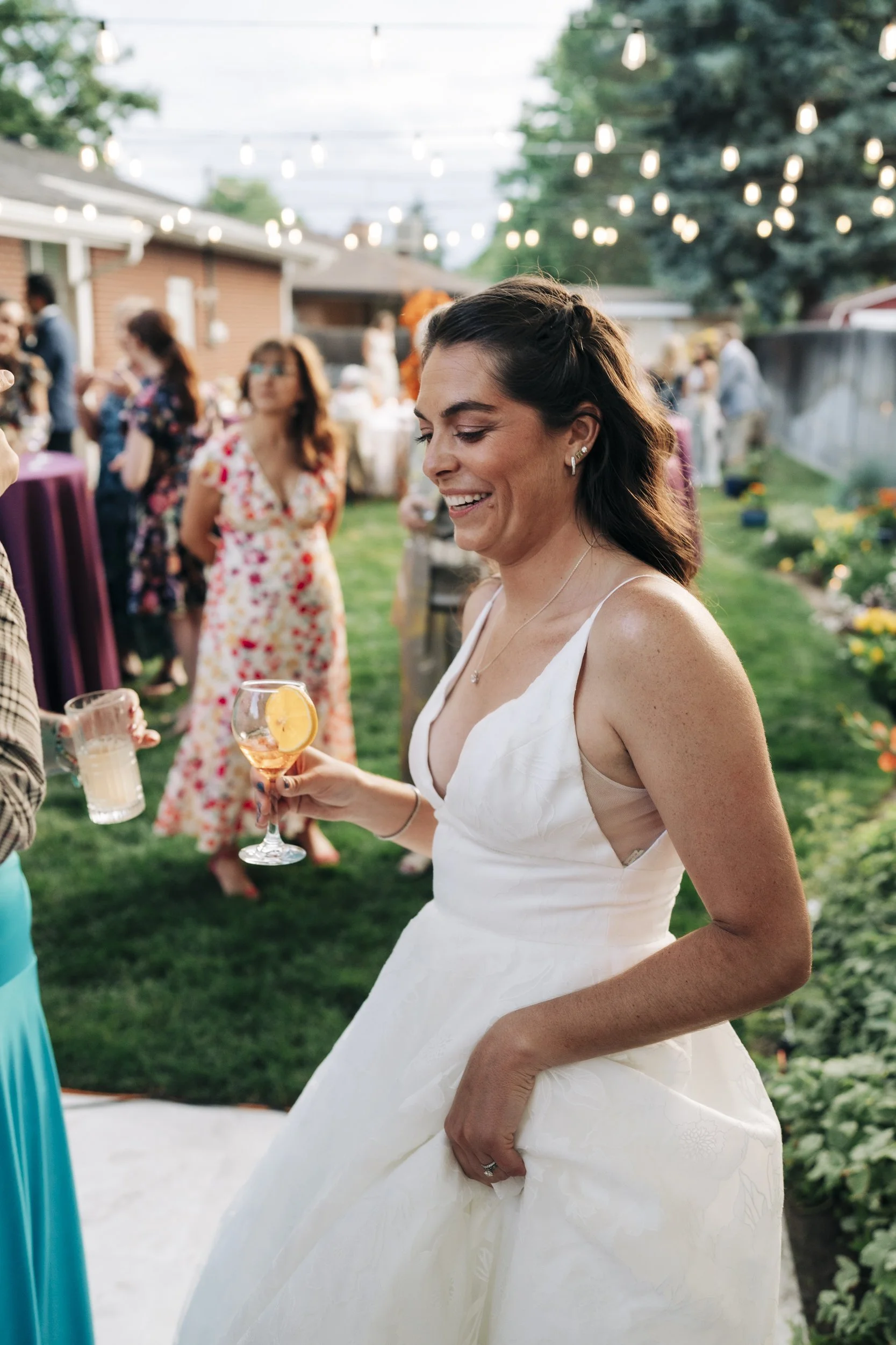 A woman in a white sleeveless wedding dress holding a wine glass with a slice of lemon at an outdoor wedding reception, smiling, with string lights and guests in the background.