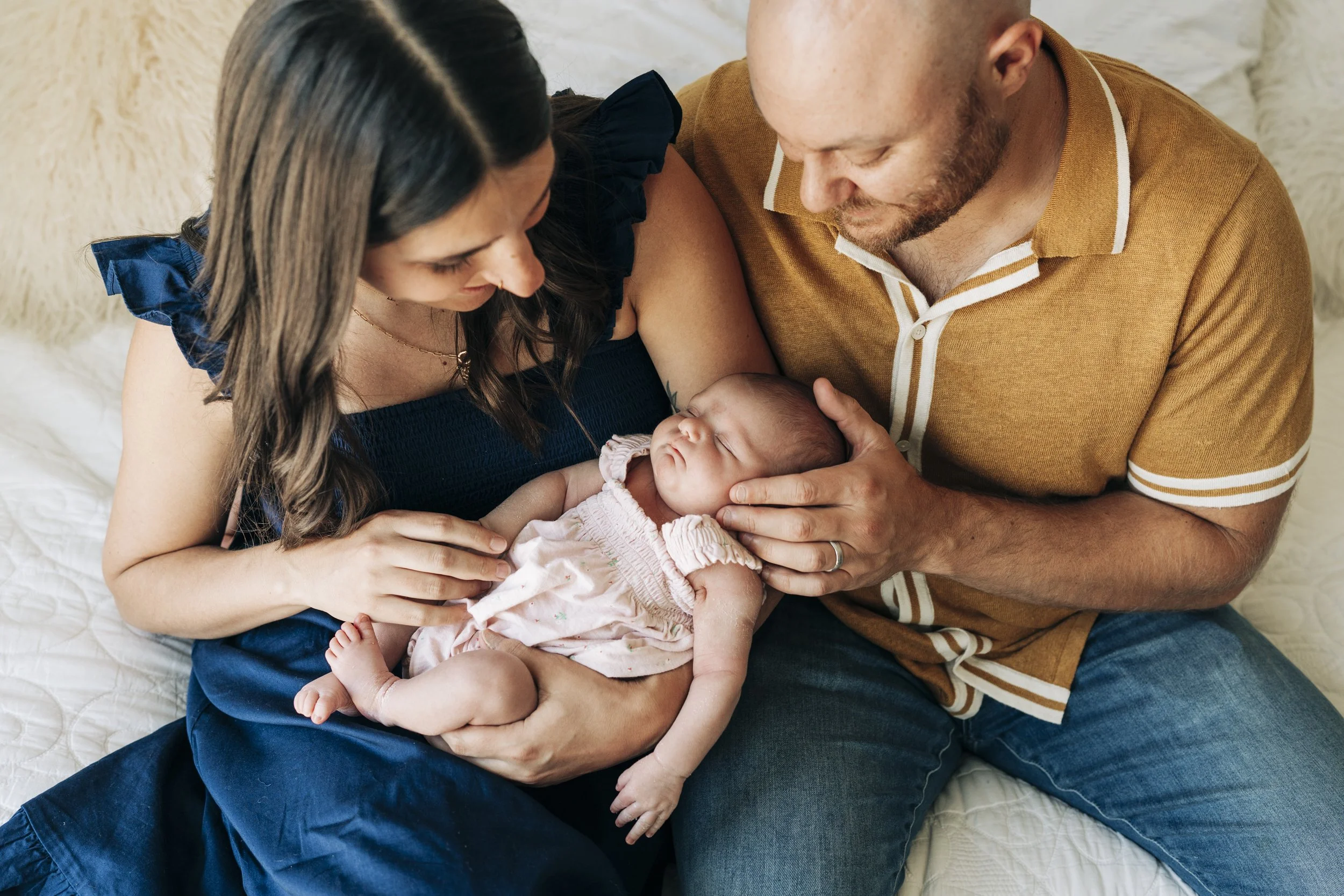 A family of three sitting on a bed, holding a sleeping newborn baby. The mother and father are looking lovingly at the baby, with the mother placing her hand on the baby's stomach and the father gently supporting the baby's head with his hand.