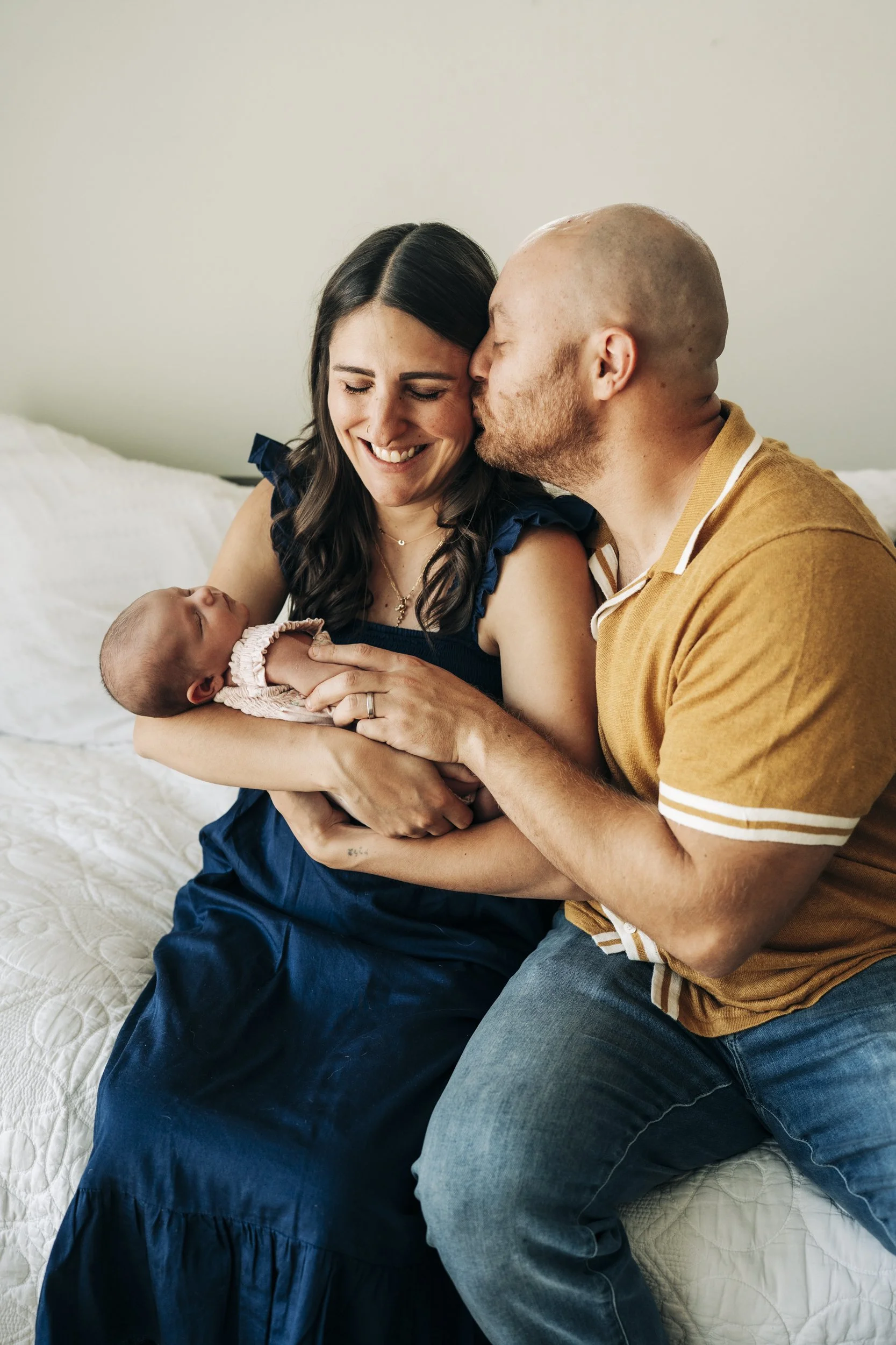 A happy family moment with a woman holding a newborn baby, a man kissing her on the cheek, and them smiling together in a bright bedroom.