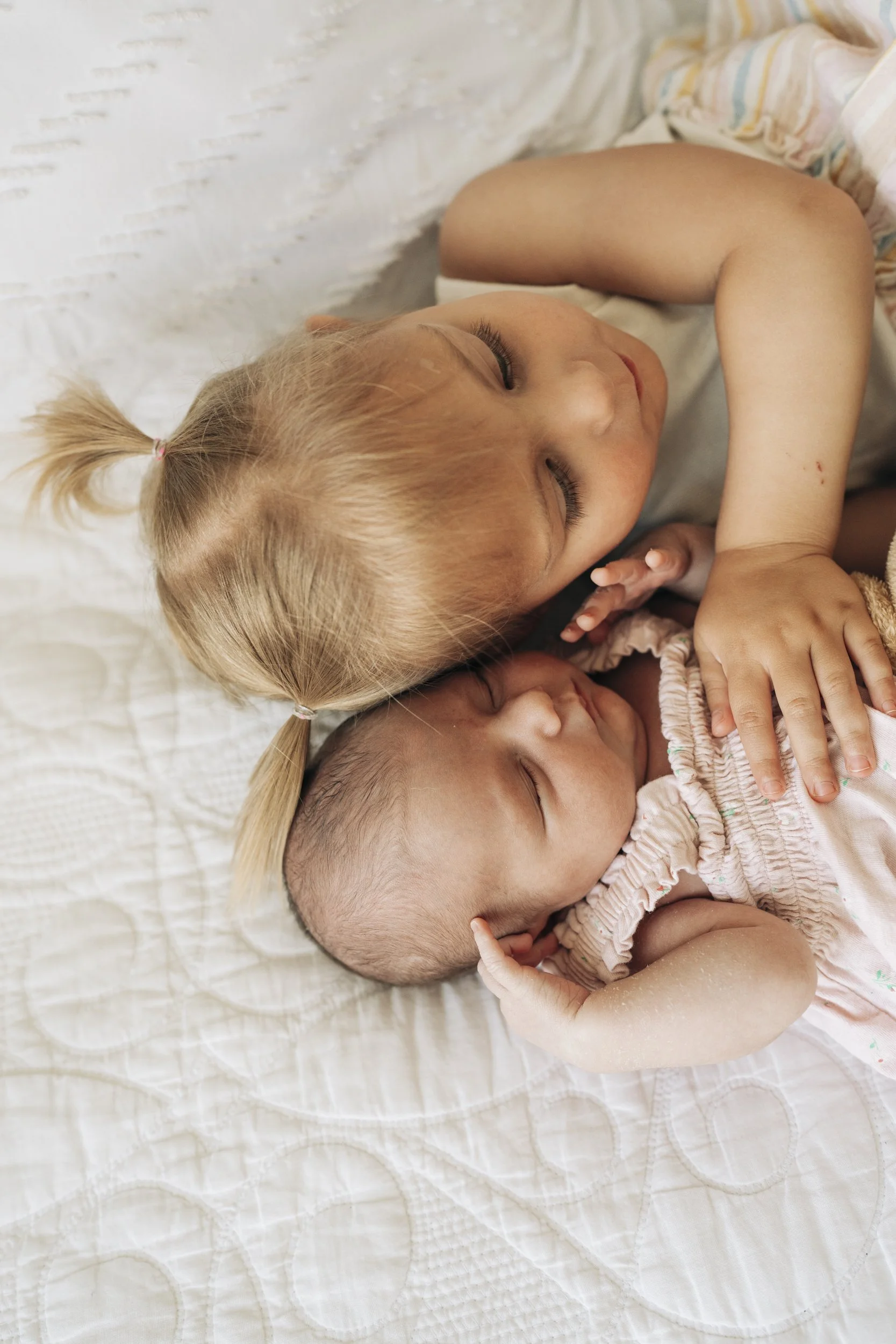 A young girl with blonde hair in pigtails is cuddling a baby with a shaved head. Both are lying on a white quilted bed, peacefully resting with eyes closed.