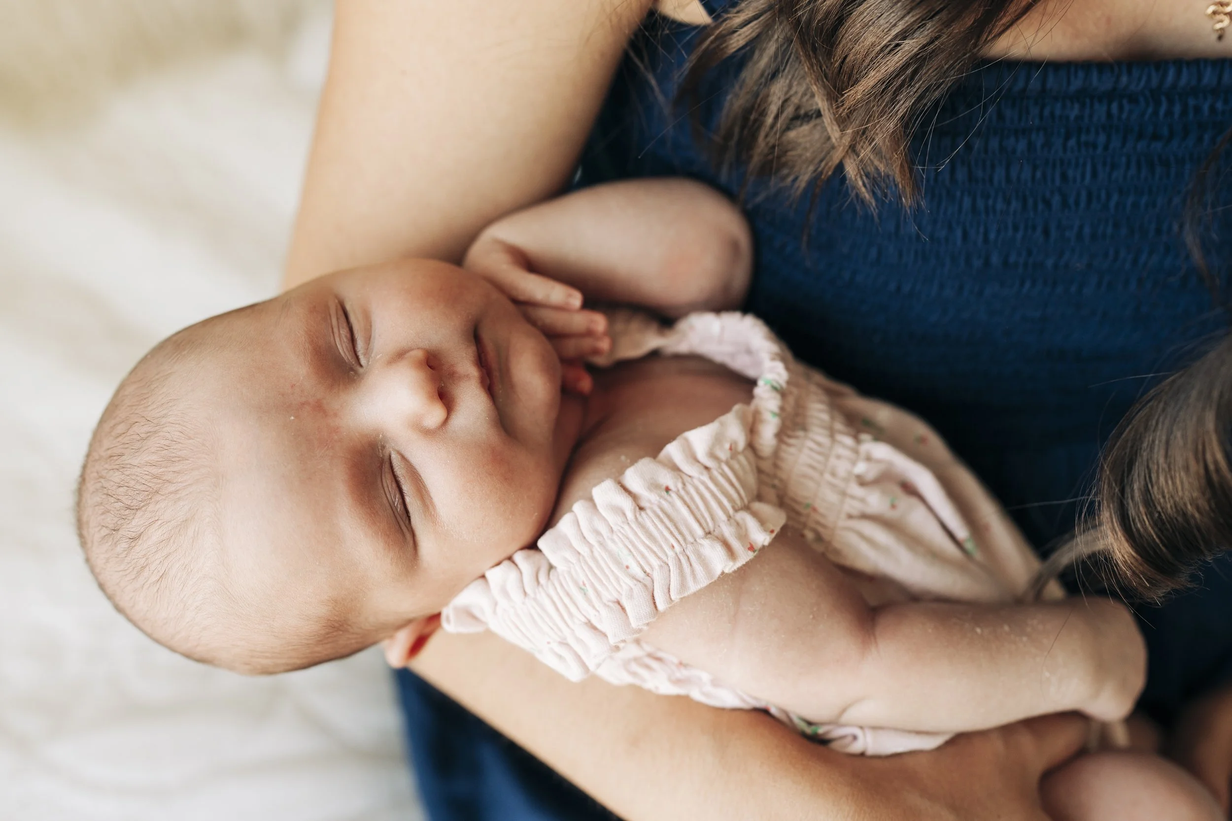 A sleeping newborn baby resting on a woman's chest, wrapped in a pink ruffled dress, while she holds the baby gently.