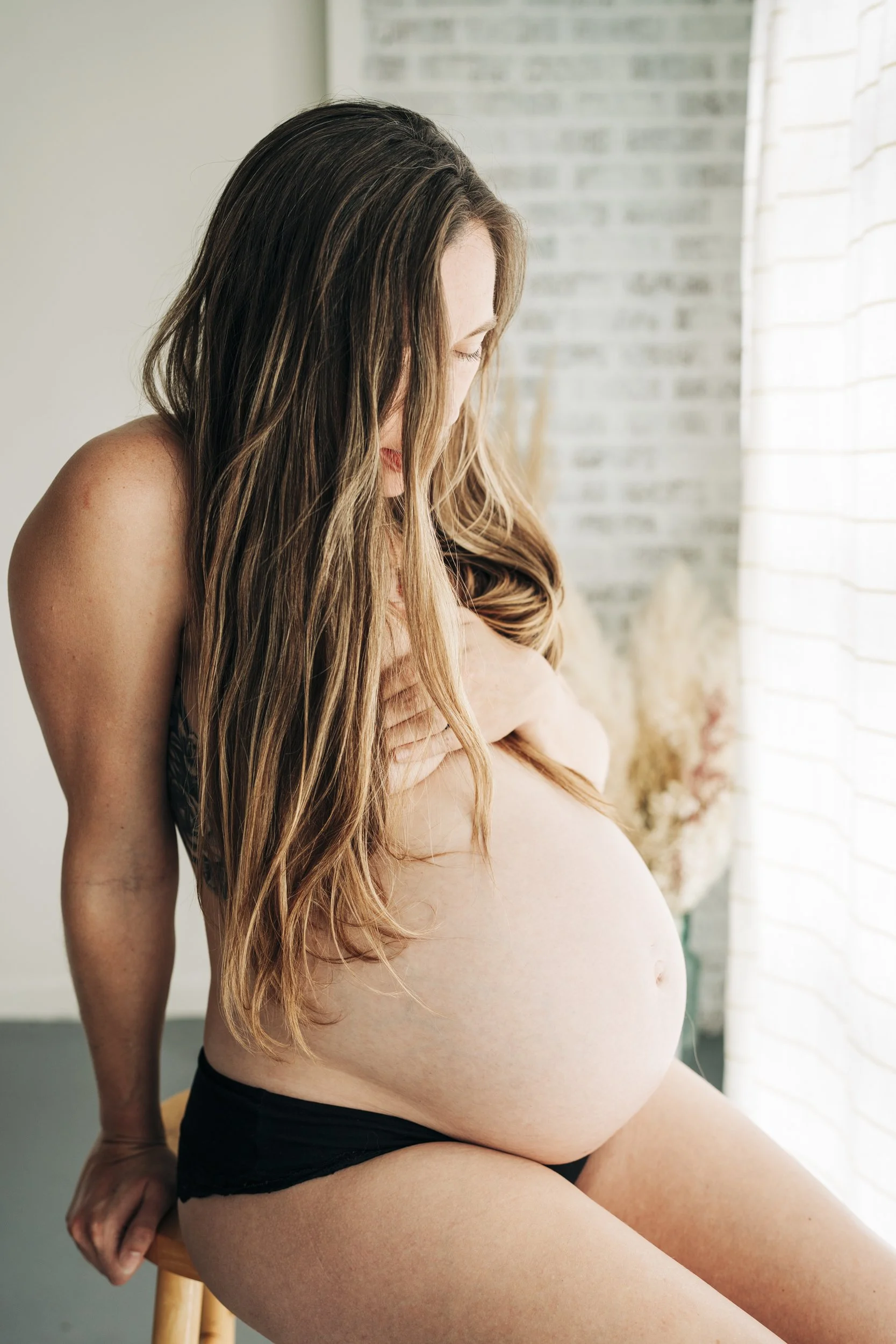 Pregnant woman with long hair sitting on a stool, lightly touching her baby bump, in a softly lit room with a white brick wall and a window with curtains.
