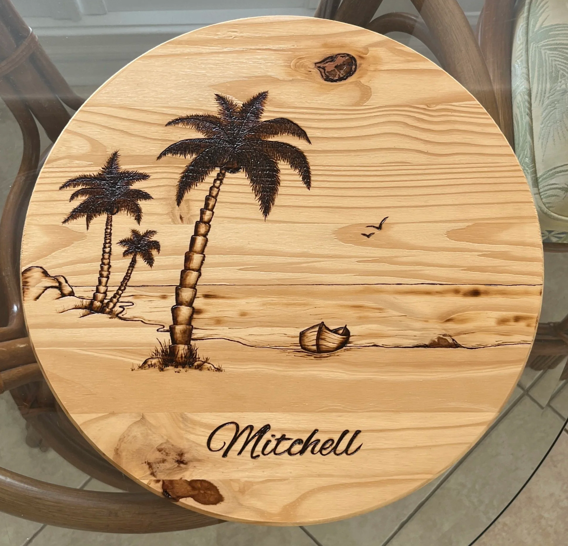 Round custom wood-burning artwork featuring a tropical beach scene with palm trees, a small sailboat, birds, and the name “Mitchell” in script lettering.