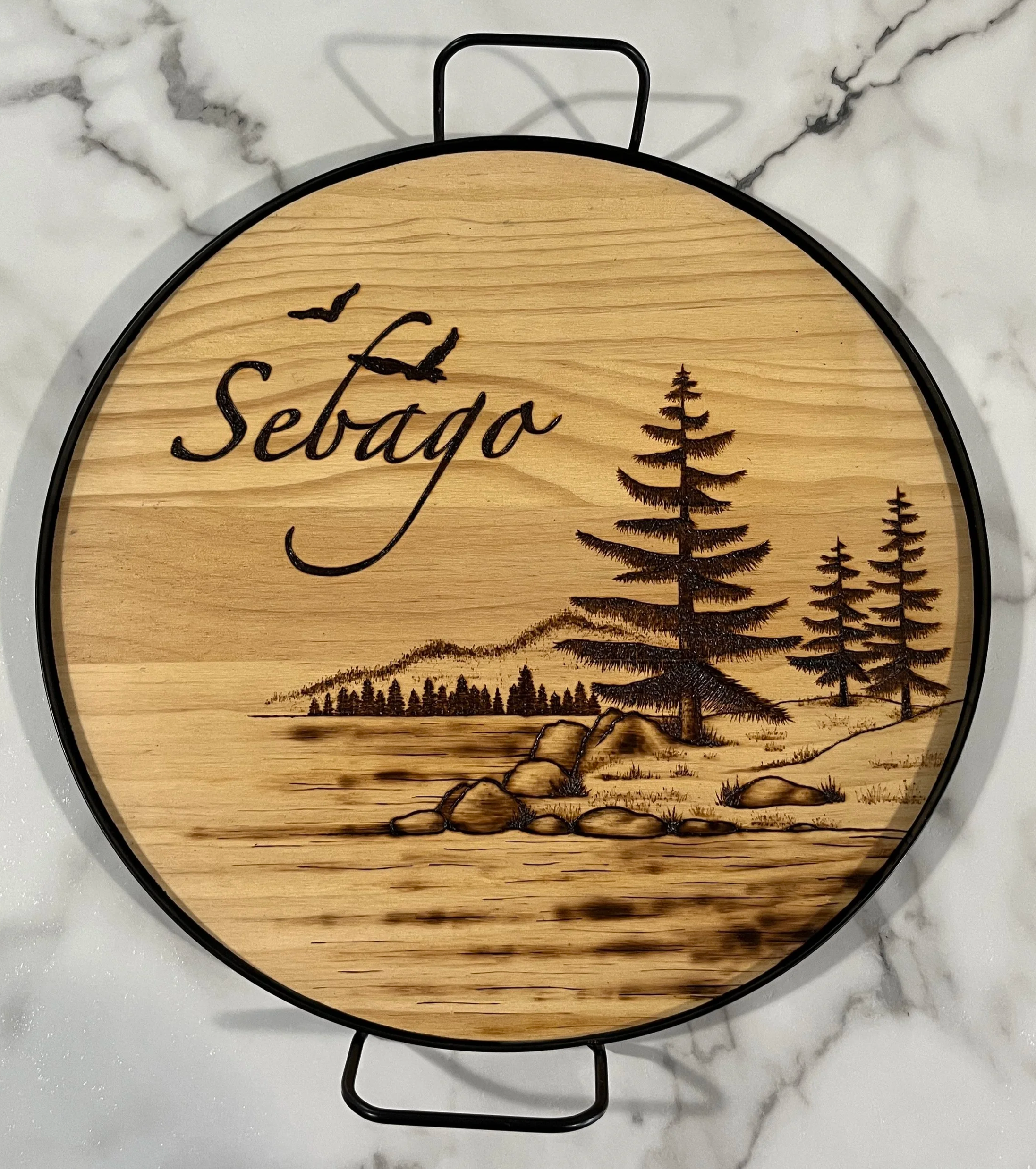 Round wood-burning artwork featuring the word “Sebago” above a lakeside landscape with pine trees, rocks, water, and flying birds.