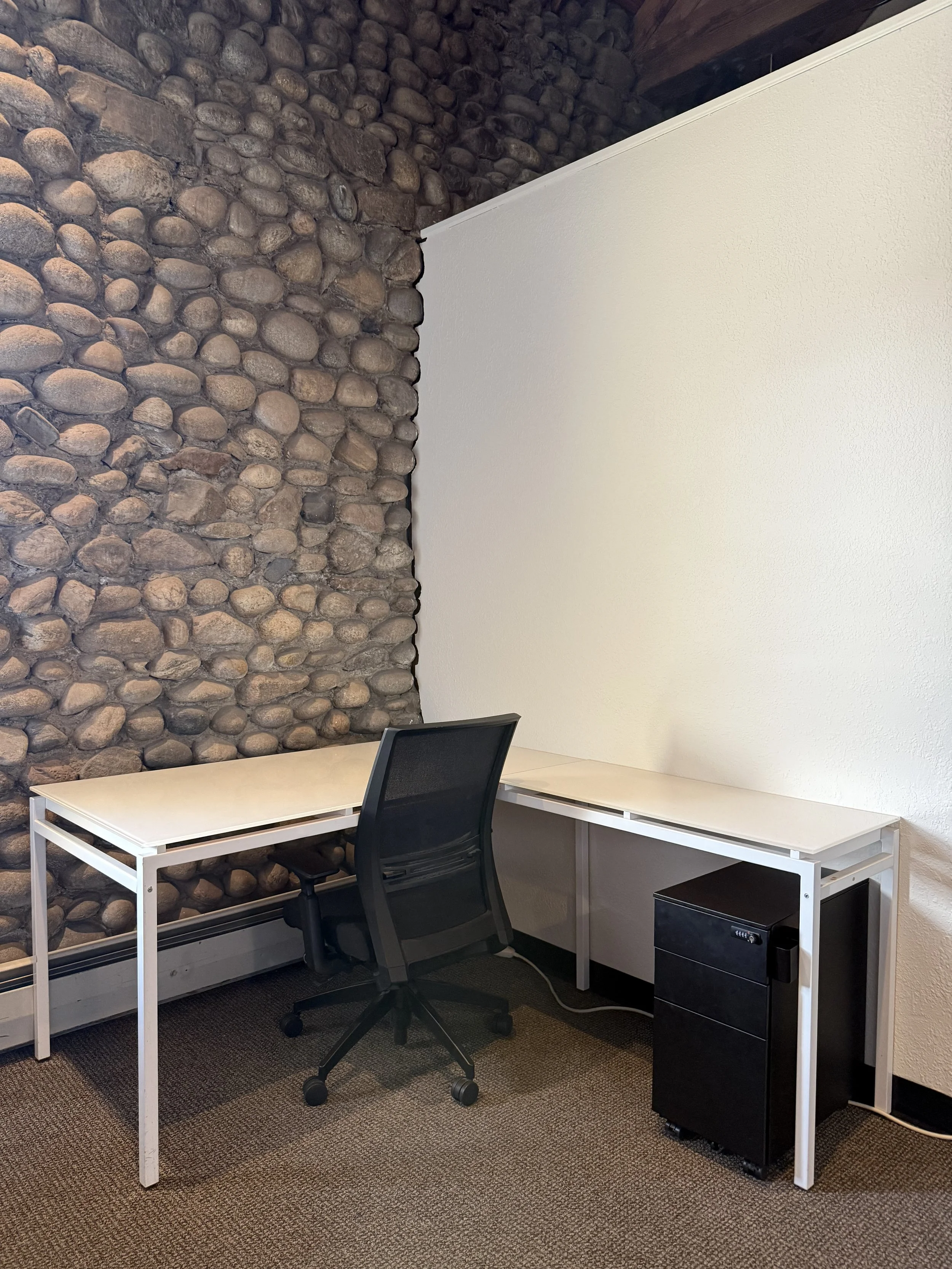 A minimalistic office space with a white desk, black office chair, a stone accent wall, and a white wall with a textured finish. There is a black computer tower on a small black filing cabinet next to the desk. The room has dark carpeting.