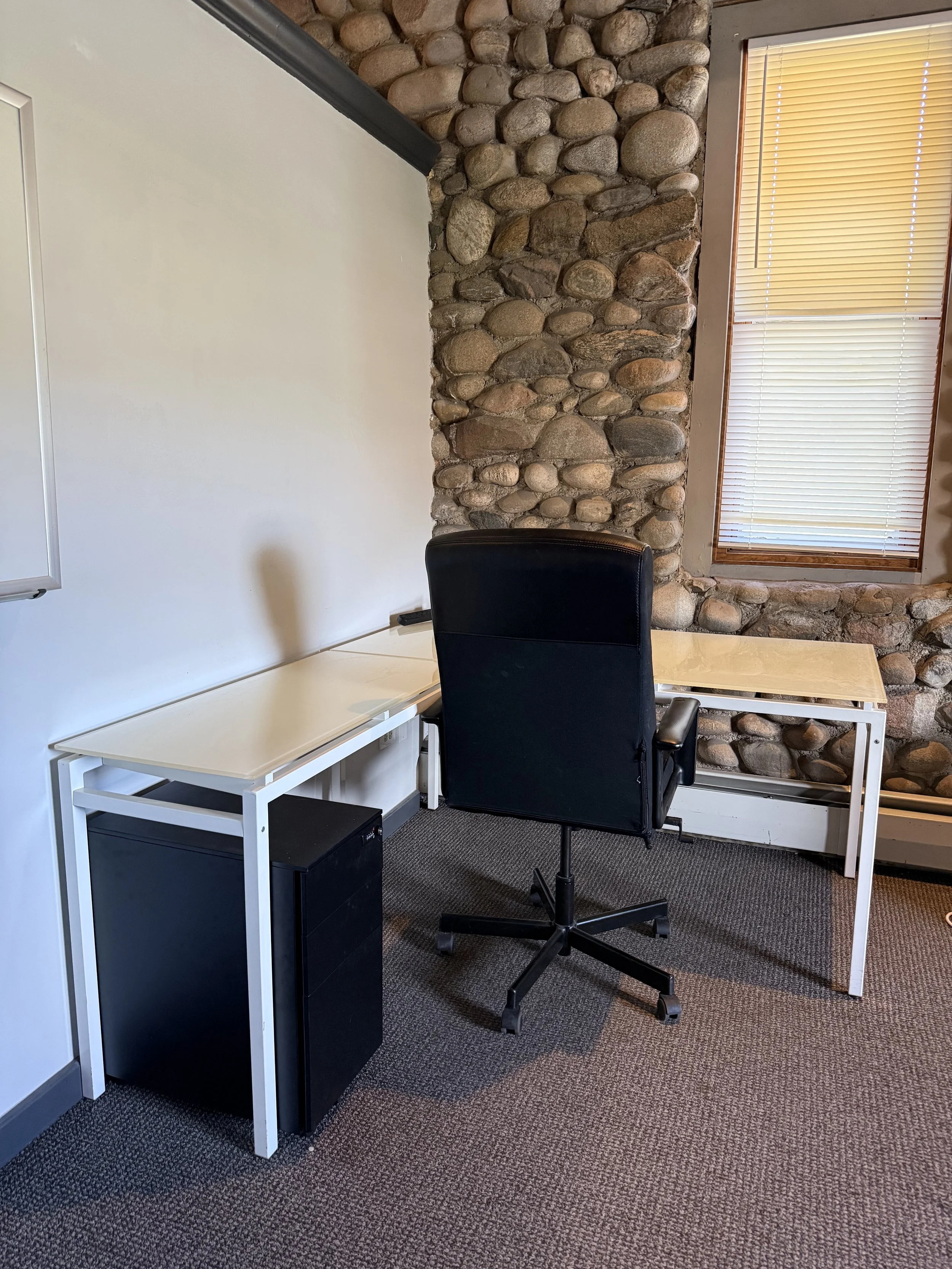 Office space with a white desk, black filing cabinet underneath, a black office chair, and a stone wall behind. There's a window with closed blinds on the stone wall, and a whiteboard on the left.