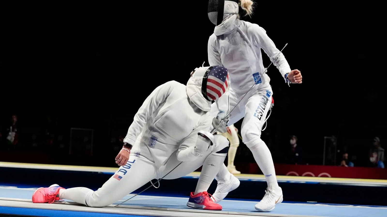 Two female American Olympic fencers in white fencing gear, with one kneeling and the other standing, on fencing piste with black background.