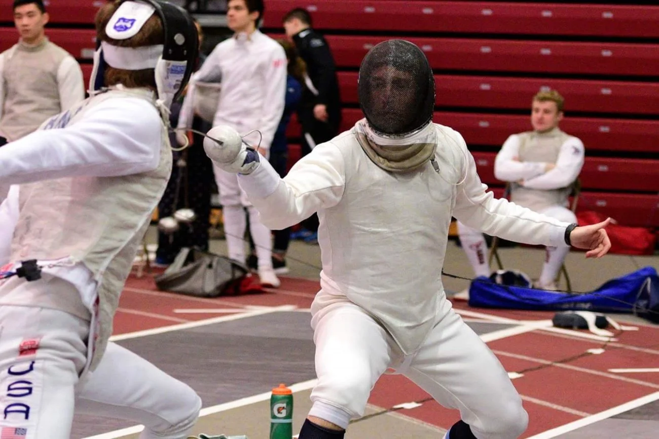 Two fencers in full gear engaging in a fencing match, with one lunging forward and the other preparing to defend, inside a gymnasium with red bleachers and several spectators in the background.