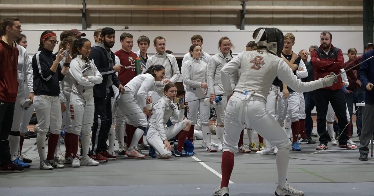 Fencer in white uniform performing a lunge while an audience of students and coaches watch in the background.