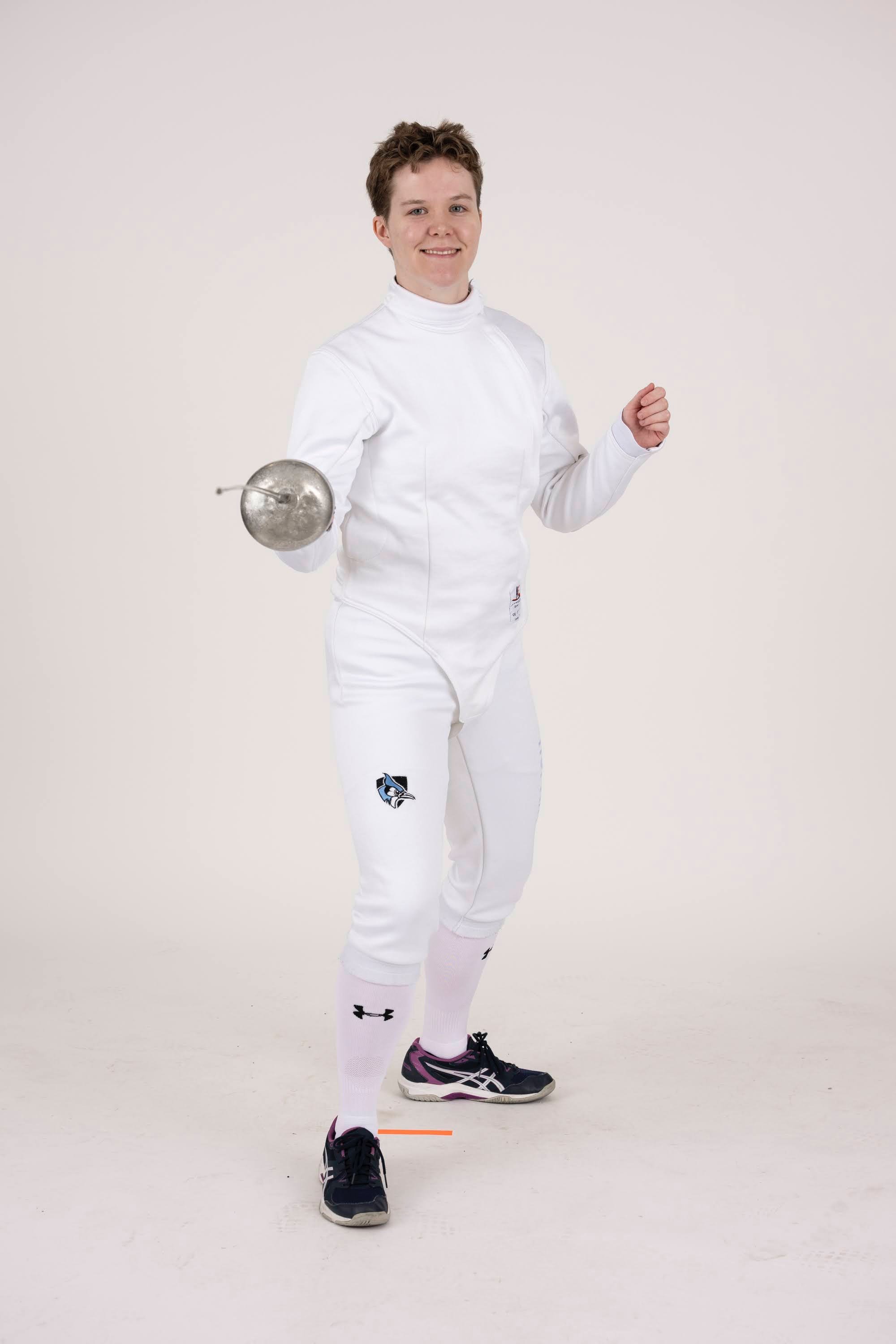 A young woman in a white fencing uniform holding a fencing foil, standing in a ready stance.