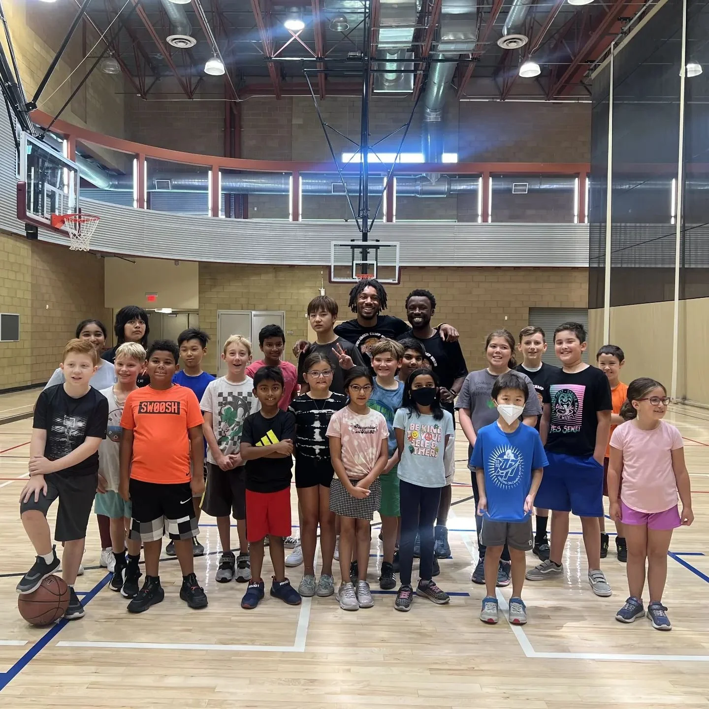 Group of children and two adult coaches posing for a photo on a basketball court inside a gymnasium, with a basketball hoop and backboard behind them.