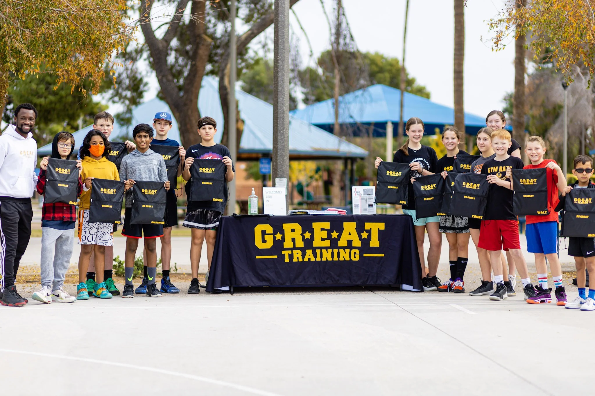 A group of children and an adult at an outdoor event, holding black drawstring bags with yellow writing, standing behind a table with a black tablecloth that says "GREAT TRAINING." The children are smiling, and some are wearing athletic clothing. The