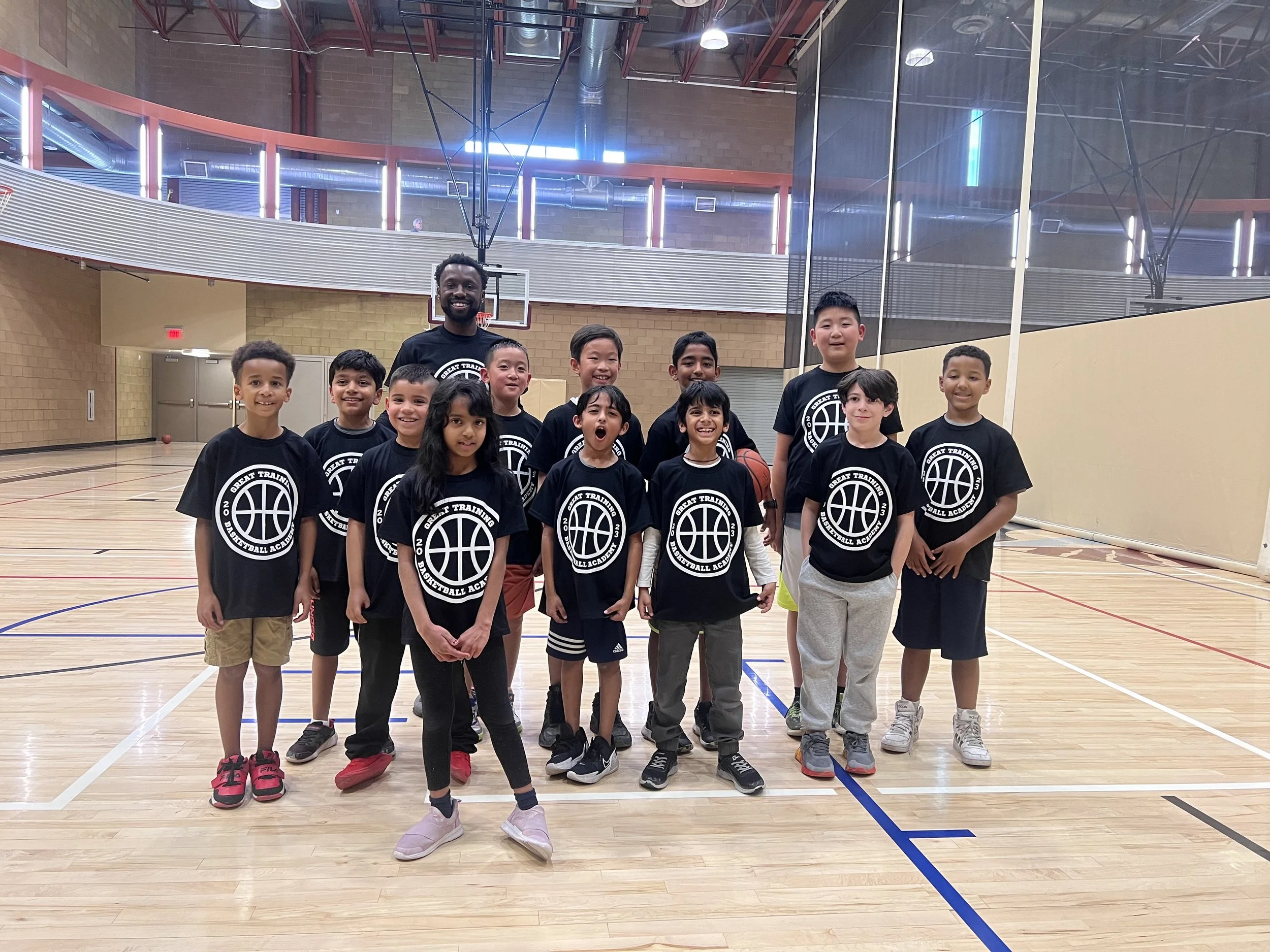Group of young boys and their coach in a basketball gym, all wearing matching black T-shirts with a basketball logo and the words 'Great Training Basketball Academy'.