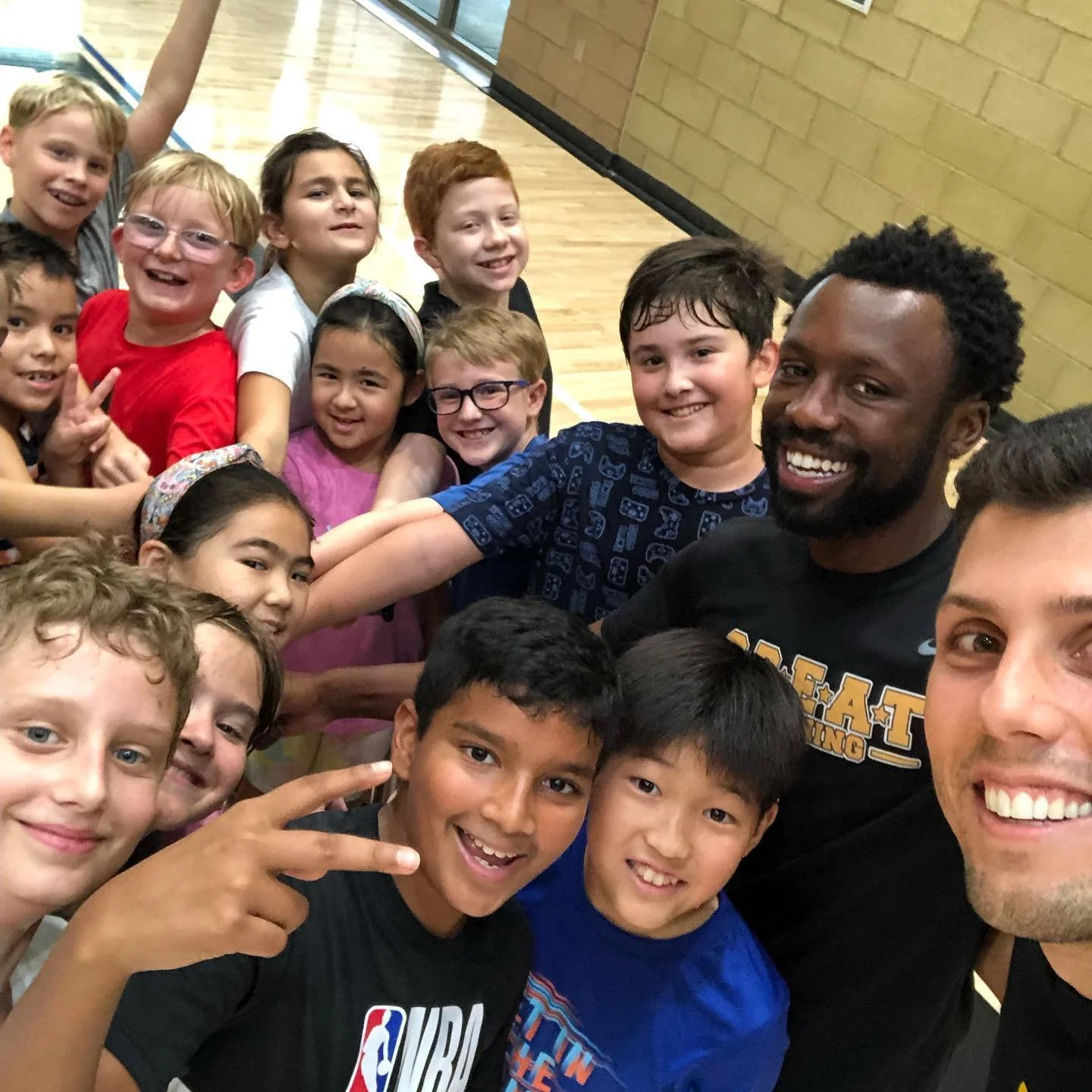 Group of children and two adults taking a group selfie in a gymnasium or school gym, all smiling and making peace signs.