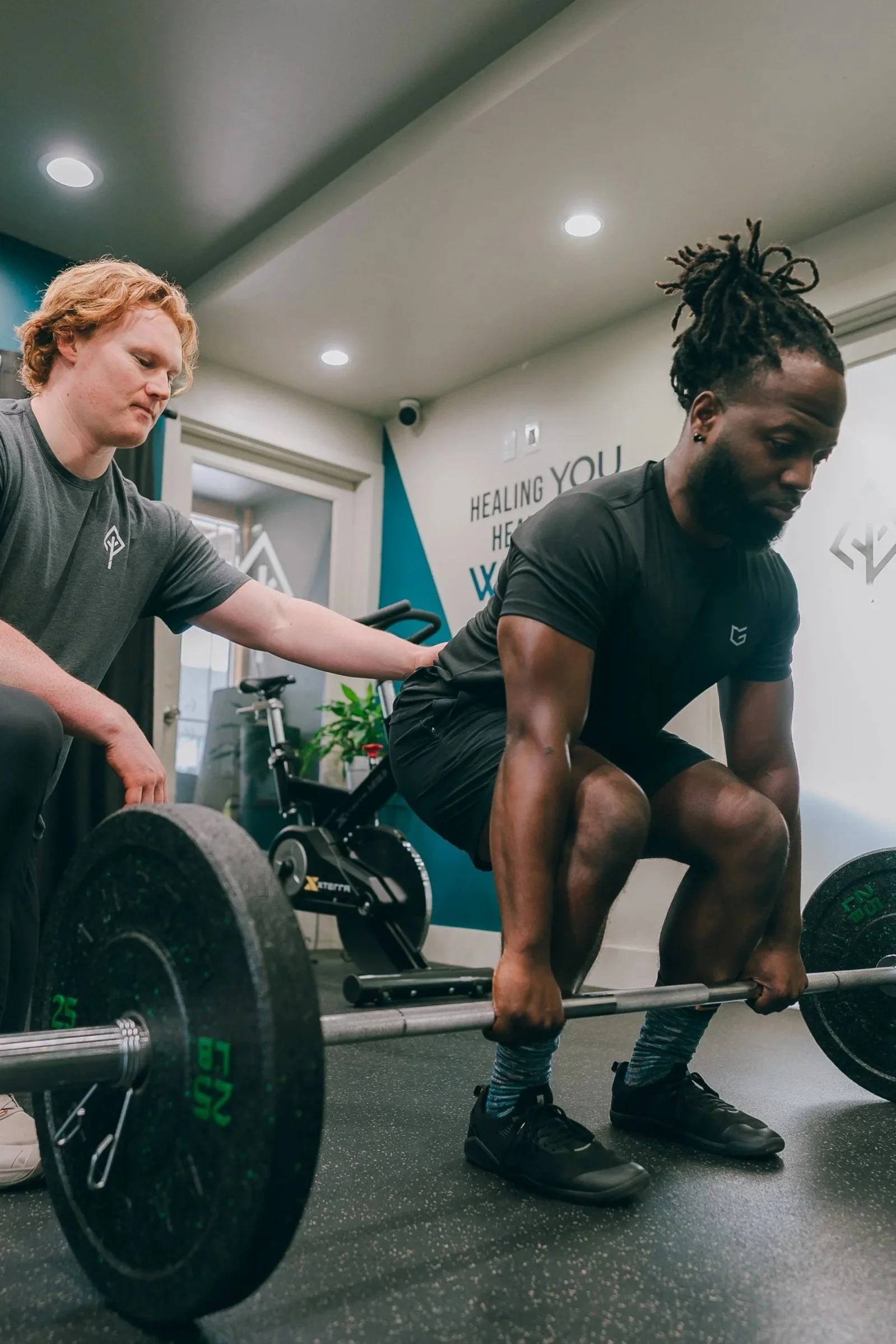 Two men in a gym, one is lifting weights, the other is spotting him and touching his shoulder