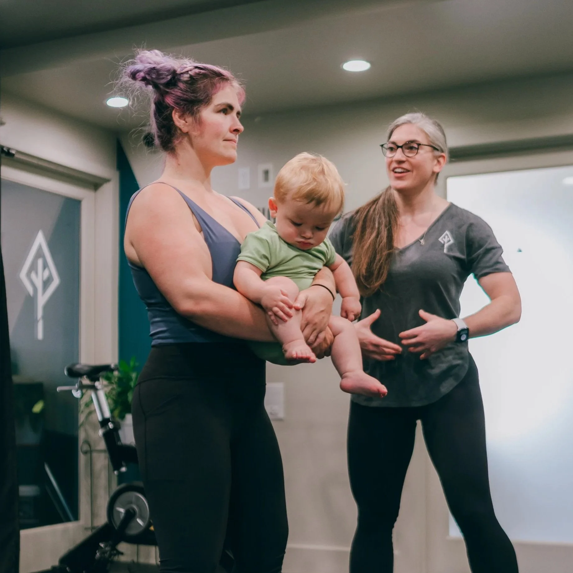 Three women and a child standing indoors, one woman is holding the child, a woman with purple hair and a woman with glasses are smiling and talking, and a bicycle is in the background.