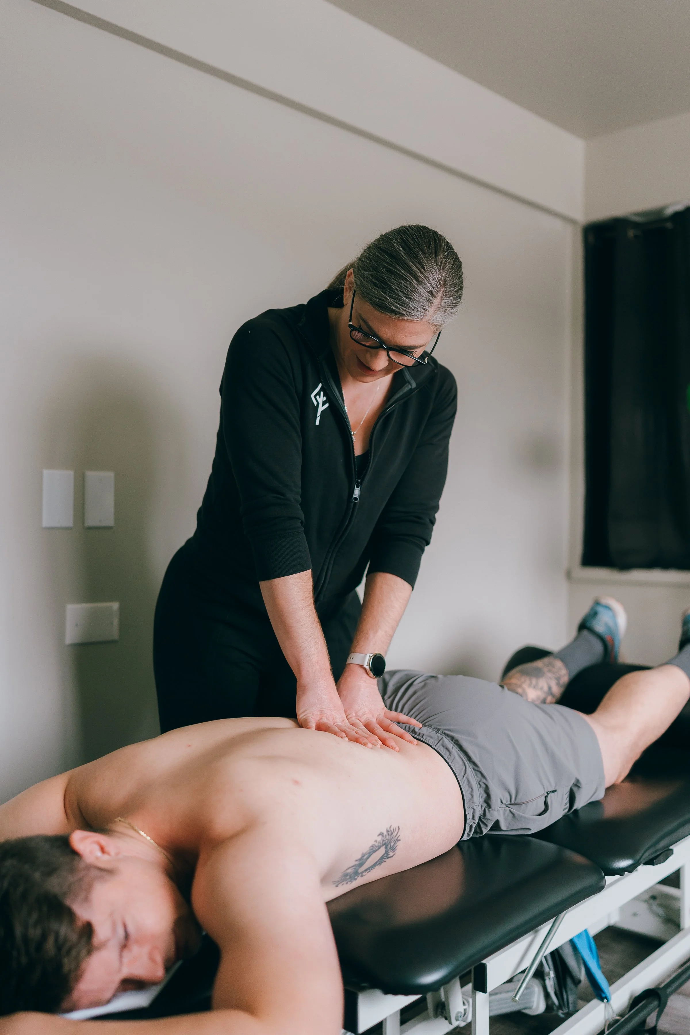A chiropractor or physiotherapist performs a spinal adjustment on a shirtless man lying face down on a treatment table, in a clinical setting with a white wall and a black curtain in the background.