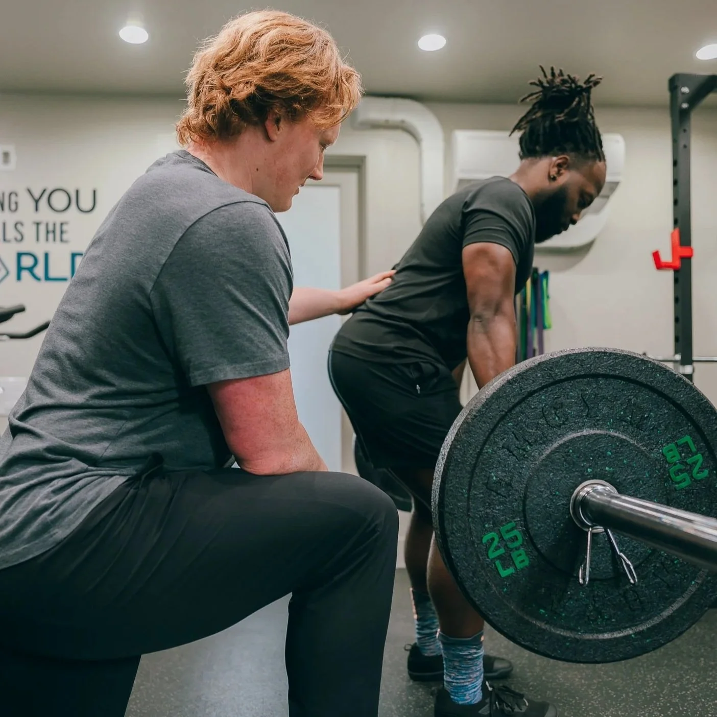 Two men in a gym, one lifting a barbell and the other spotting him, with a focus on their upper bodies.