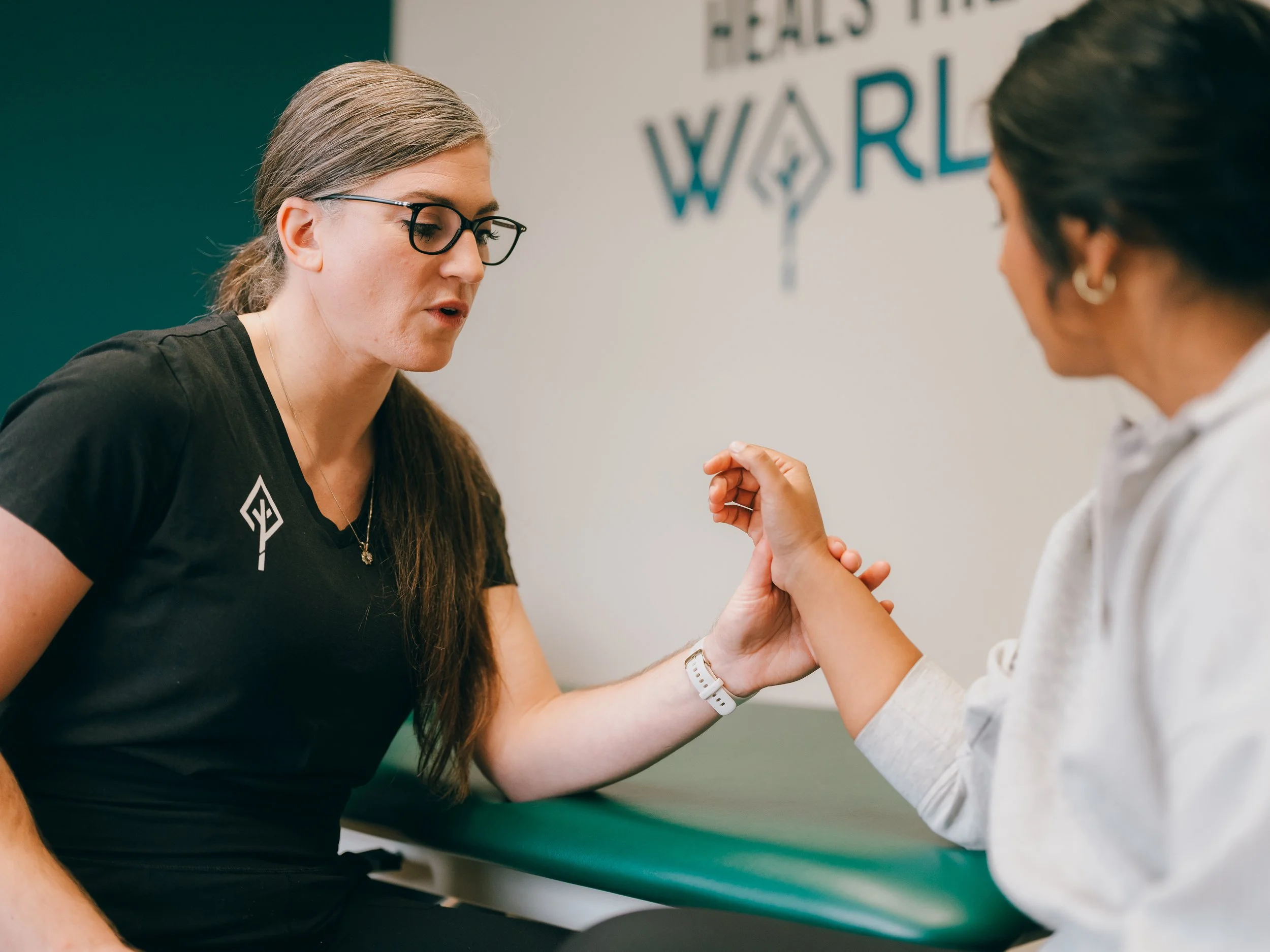 A woman with glasses and long hair is arm wrestling with another woman whose face is not visible. They are sitting at a table in a room with a blurred logo in the background.