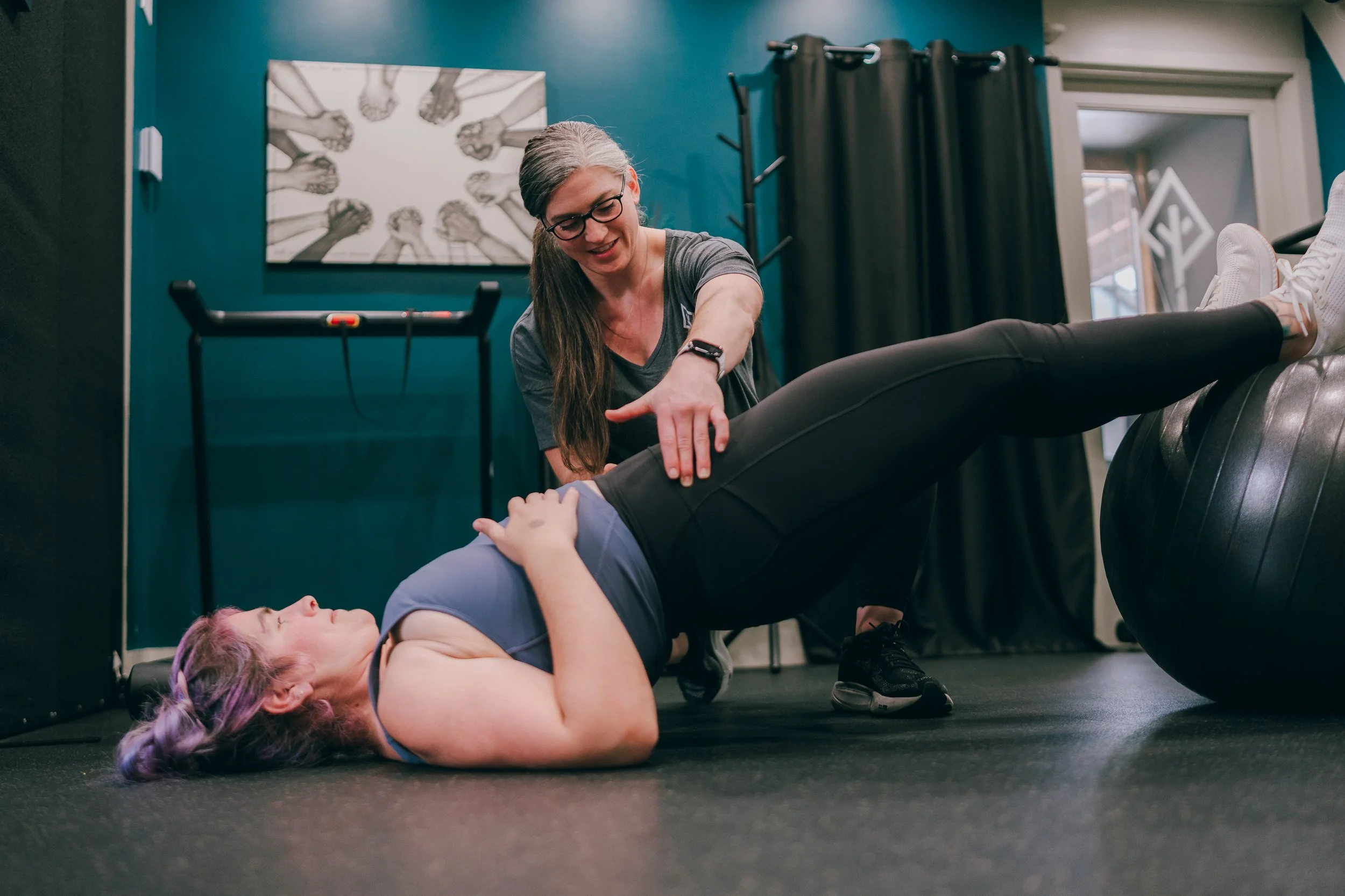 A woman with gray hair and glasses helping a person with purple hair on the floor during a fitness or rehabilitation session in a gym or physical therapy center.