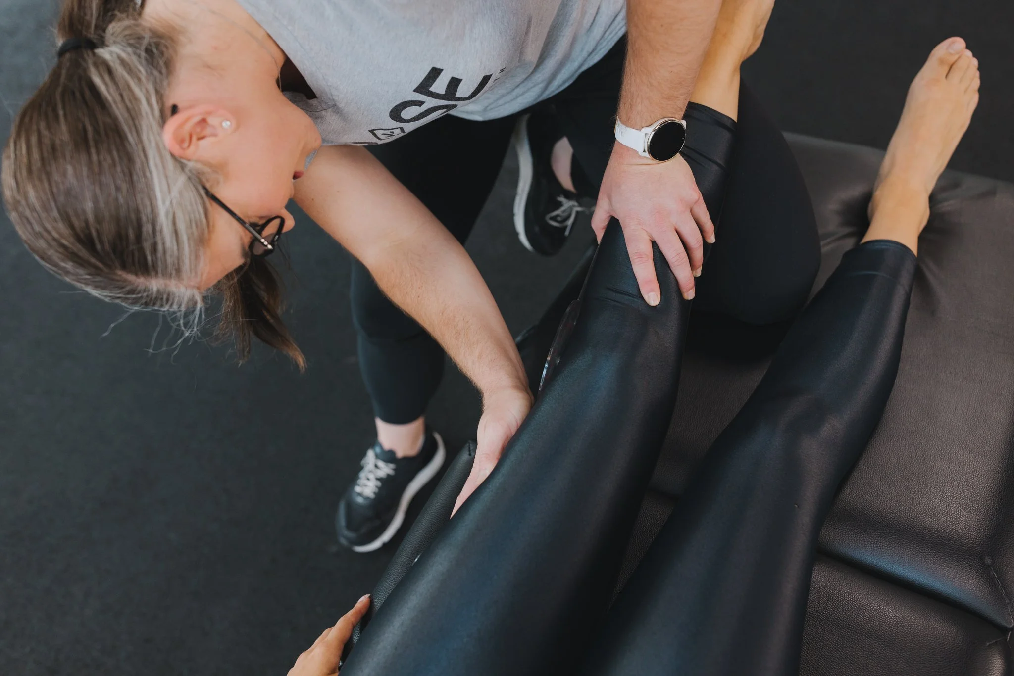 A physical therapist or trainer assisting a woman with stretching or exercises on a black padded table.