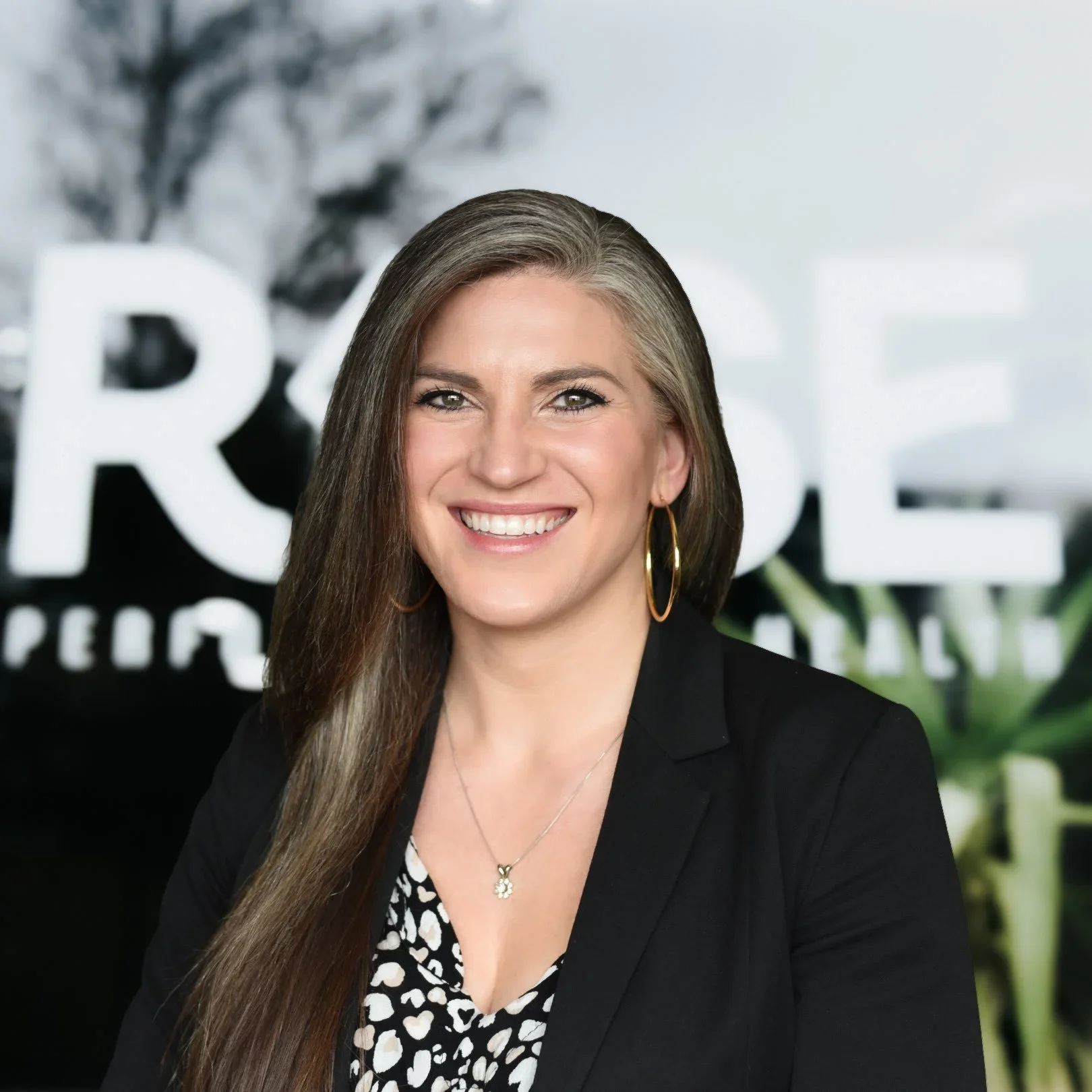 Close-up of a smiling woman with long brown hair and blue eyes, wearing a floral dress and earrings, outdoors with blurred green background.