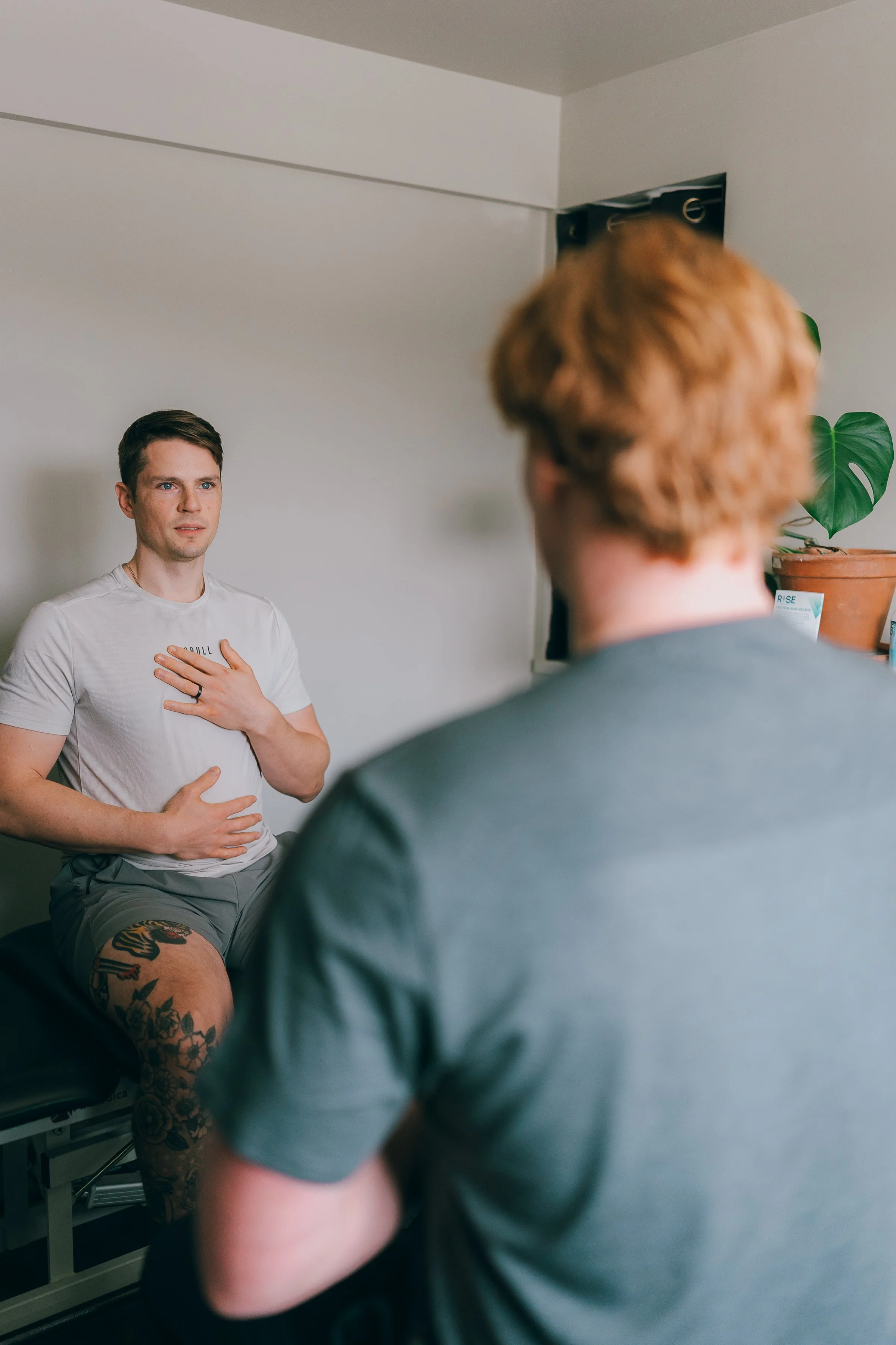 A man with a tattooed leg, sitting on a medical examination table, is doing a breath exercise or story, with one hand on his chest and the other on his stomach. He is facing a healthcare professional in a clinic or office setting.