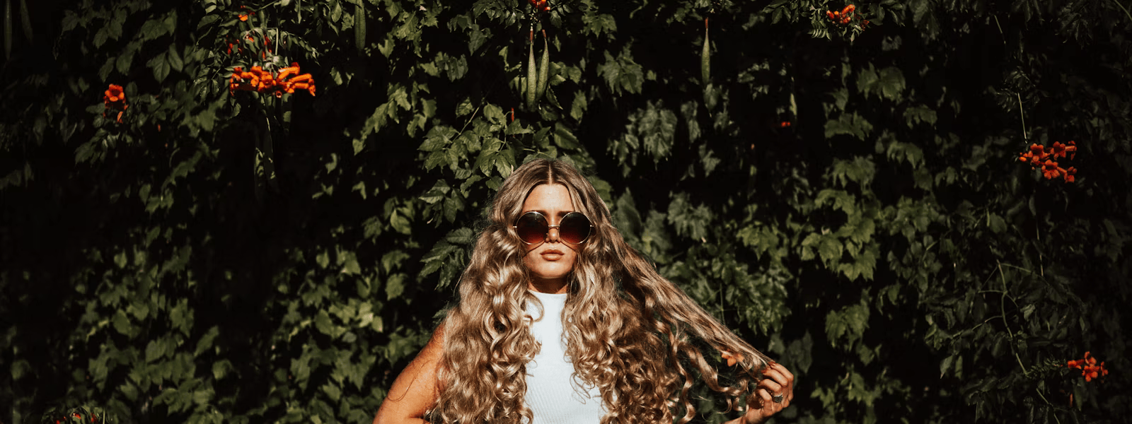 Woman with long, curly blonde hair extensions standing in front of greenery, showcasing natural volume in Portland