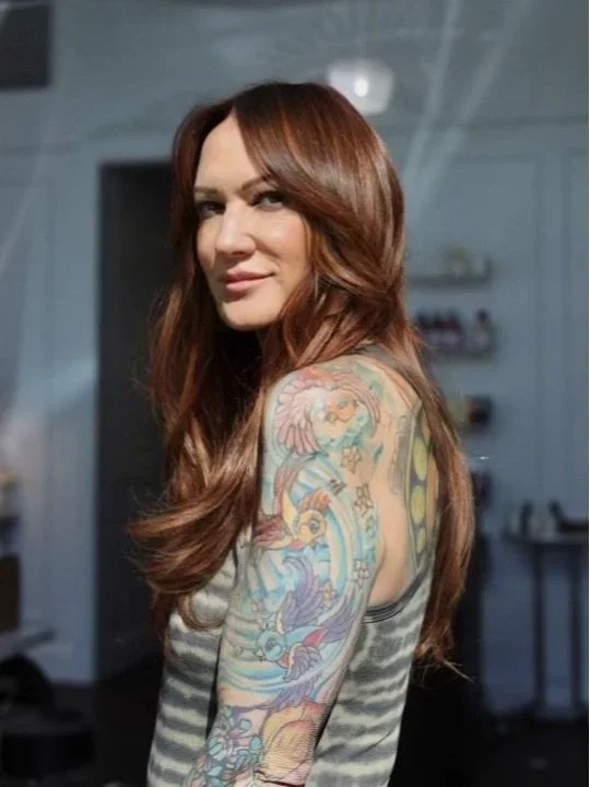Woman with long, wavy, auburn hair, visible tattoo sleeve on her right arm, wearing a sleeveless, striped top in a room with a blurred background.