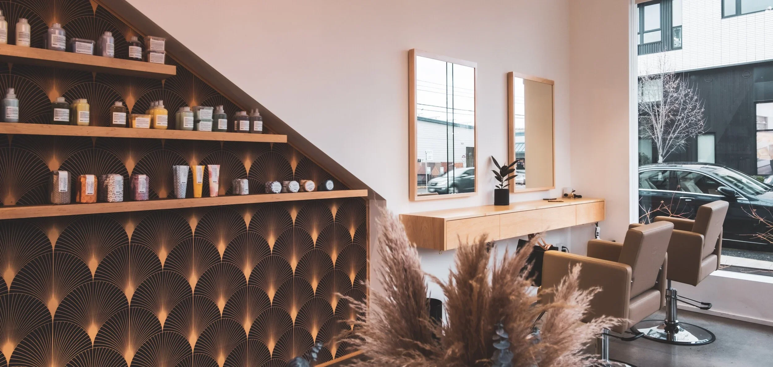 A cozy hair salon interior with a black and gold patterned accent wall, wooden shelves filled with hair products, beige salon chairs, wall mirrors, and a large window showing parked cars outside.