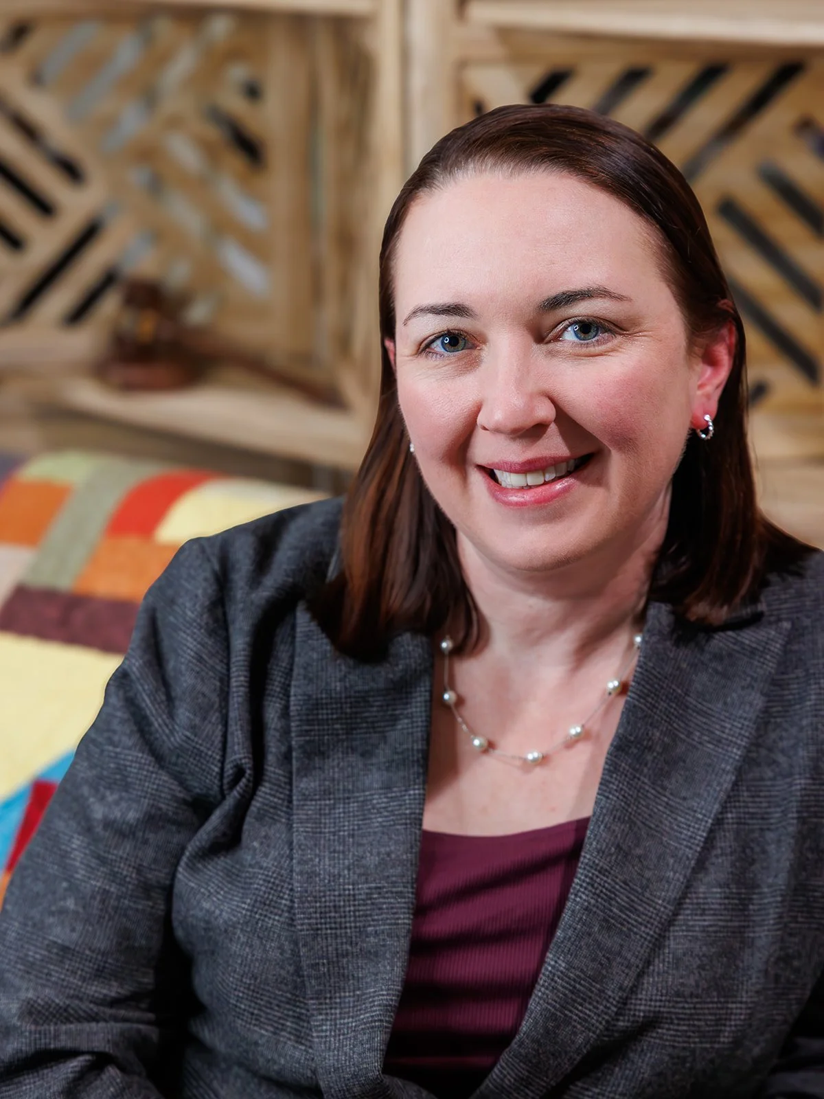 A woman with shoulder-length brown hair and blue eyes smiling at the camera, wearing a black blazer, a maroon top, and pearl jewelry, sitting in front of a wooden background and colorful quilted pillow.