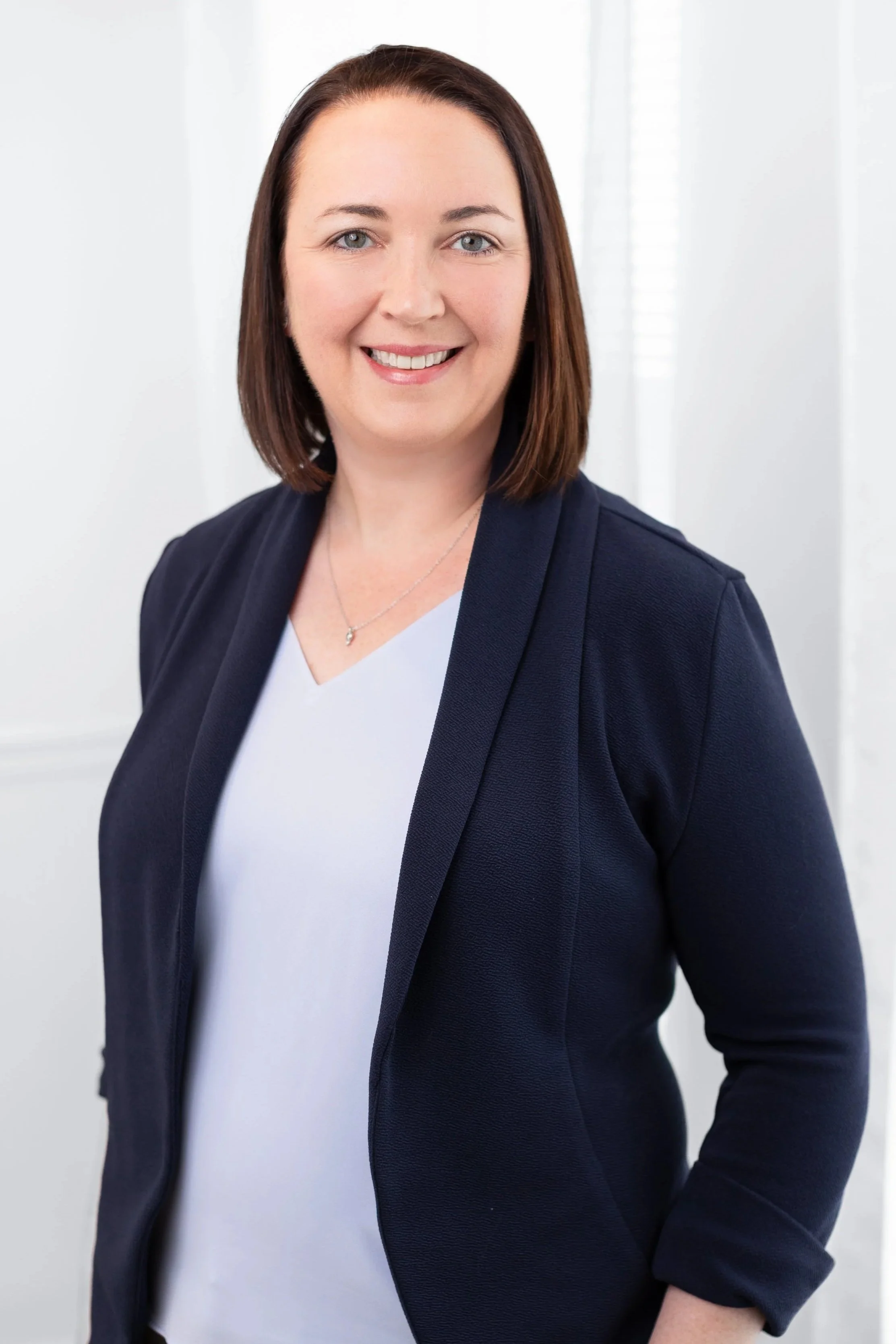 A professional woman with shoulder-length brown hair, wearing a navy blazer over a white top, smiling in front of a light background.