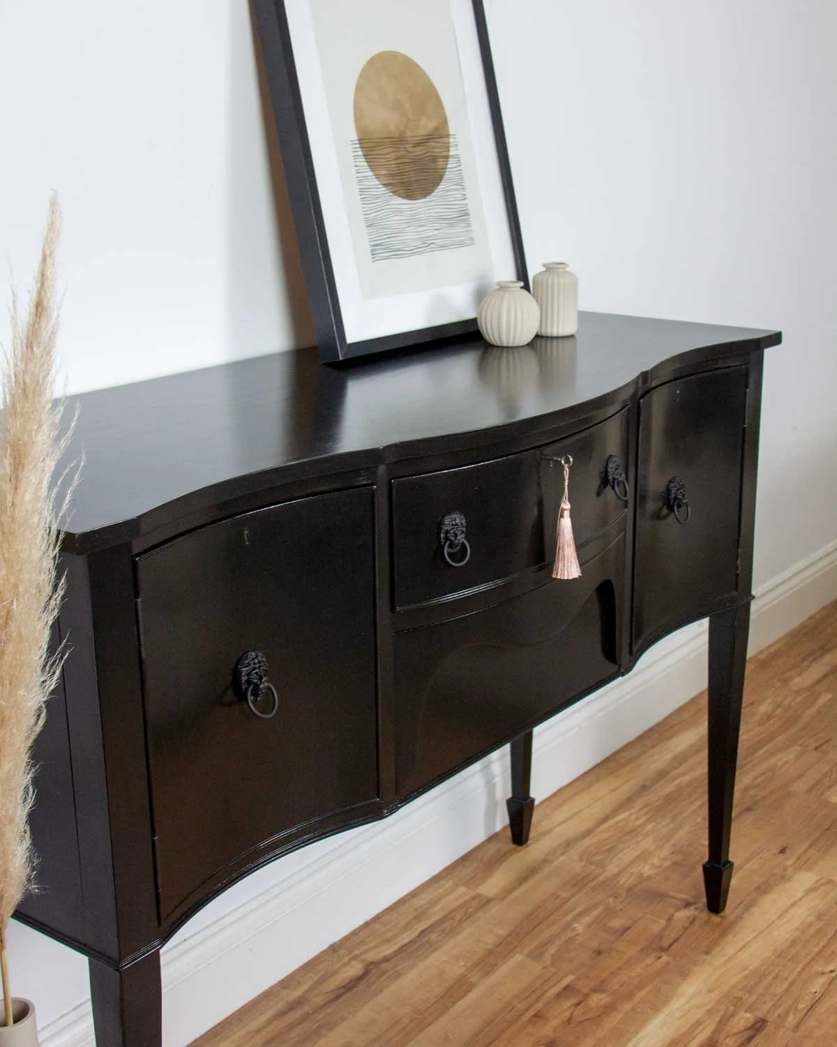 A black vintage-style sideboard with curved drawer and door fronts, decorated with framed abstract artwork and small vases on top, set against a white wall on a hardwood floor.