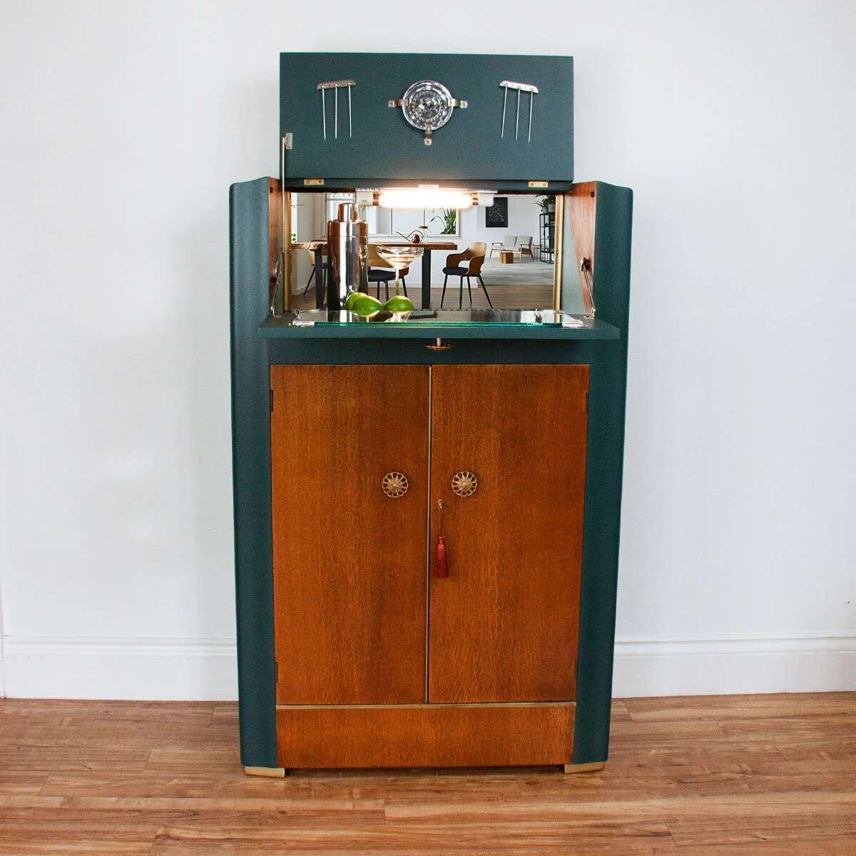 Mid-century modern wooden and green bar cabinet with mirror, metal accents, and a few limes on top, against a white wall with a wooden floor.