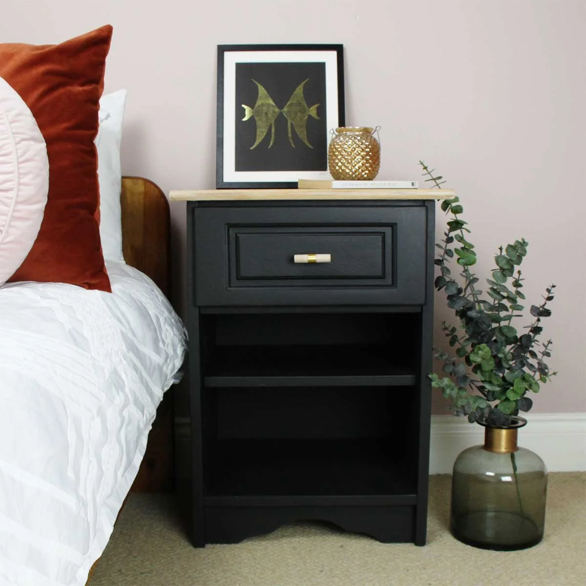 A black bedside table with a gold and white handle, decorated with a framed artwork of a fish, a gold textured vase, and a stack of books. To the right, a large glass vase with eucalyptus plant. Part of a bed with white bedding and a rust-colored pil