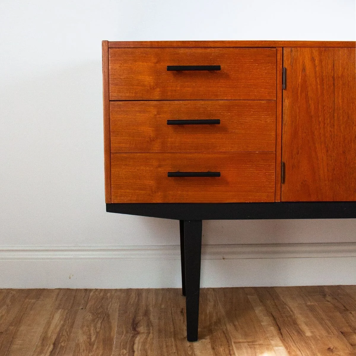 A mid-century modern wooden sideboard with three drawers and a cabinet on the right, standing against a white wall and on a wood floor.