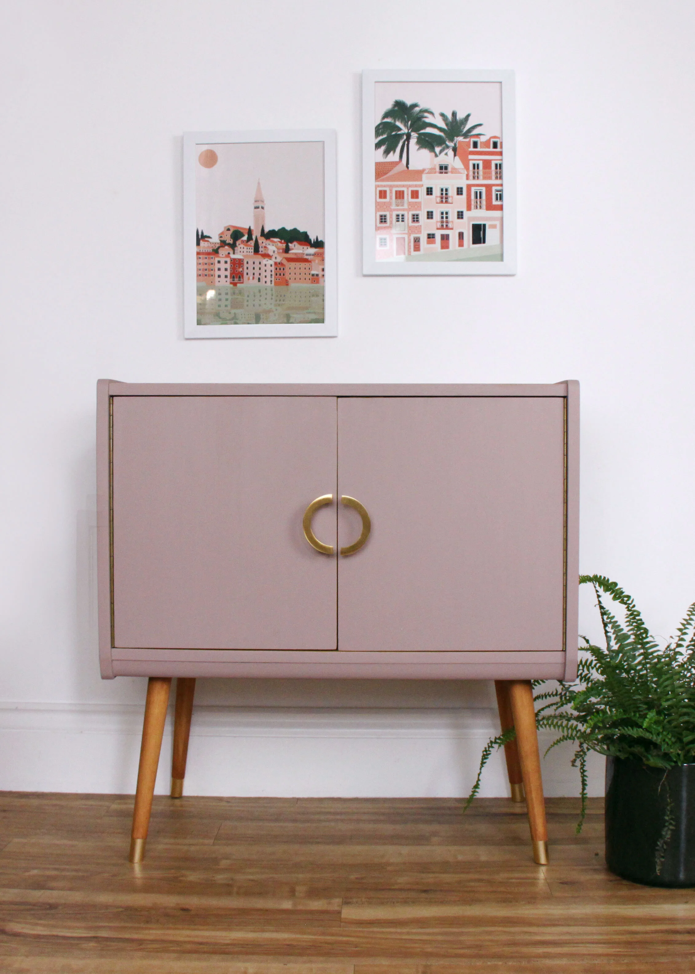 A pink mid-century modern cabinet with curved gold handles and wooden legs, placed against a white wall with two framed pictures above it. There is a potted fern on the right side of the cabinet.
