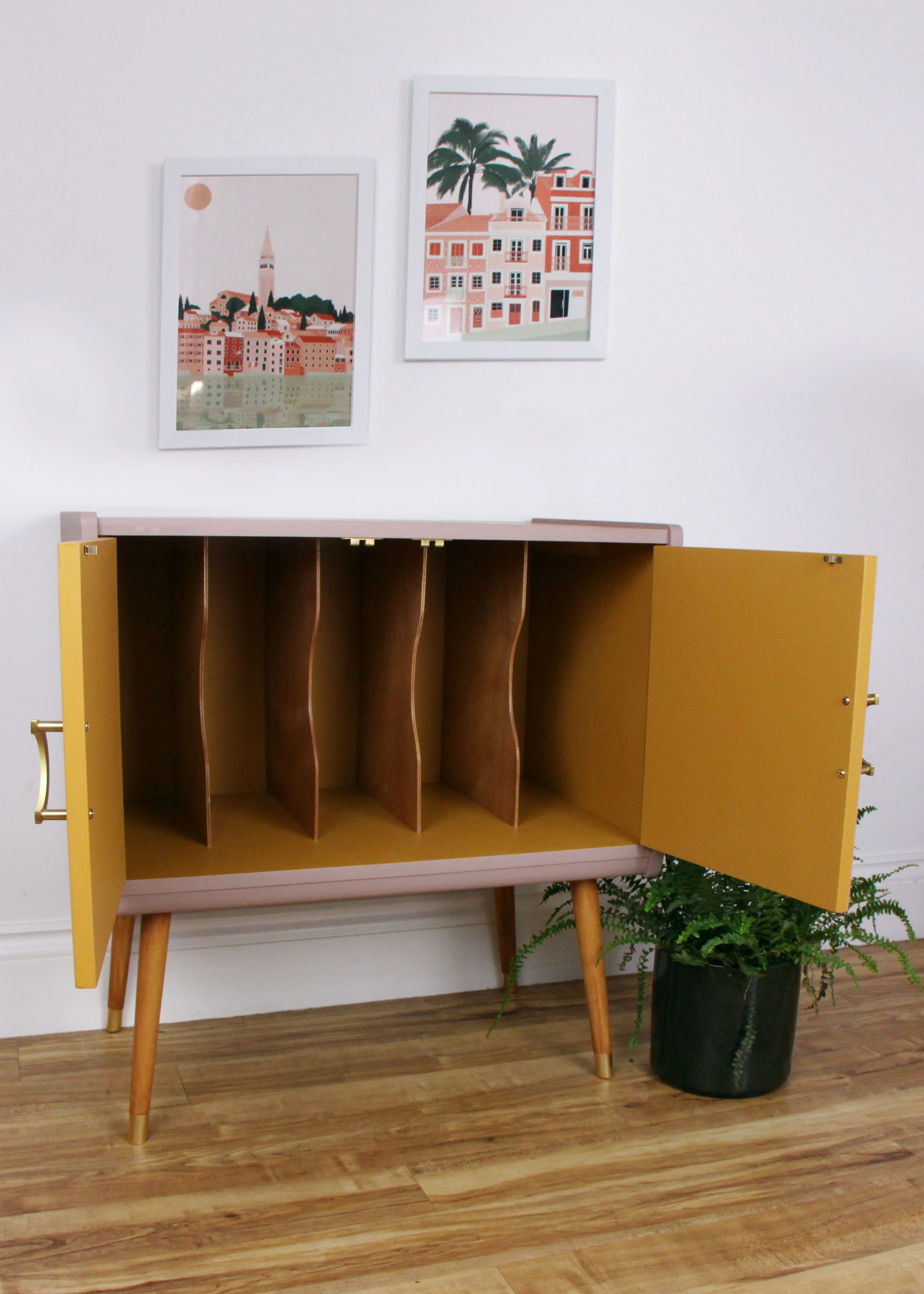 Open vintage cabinet with yellow doors and wooden compartments, next to a potted fern plant, with two framed cityscape art prints on the white wall above.