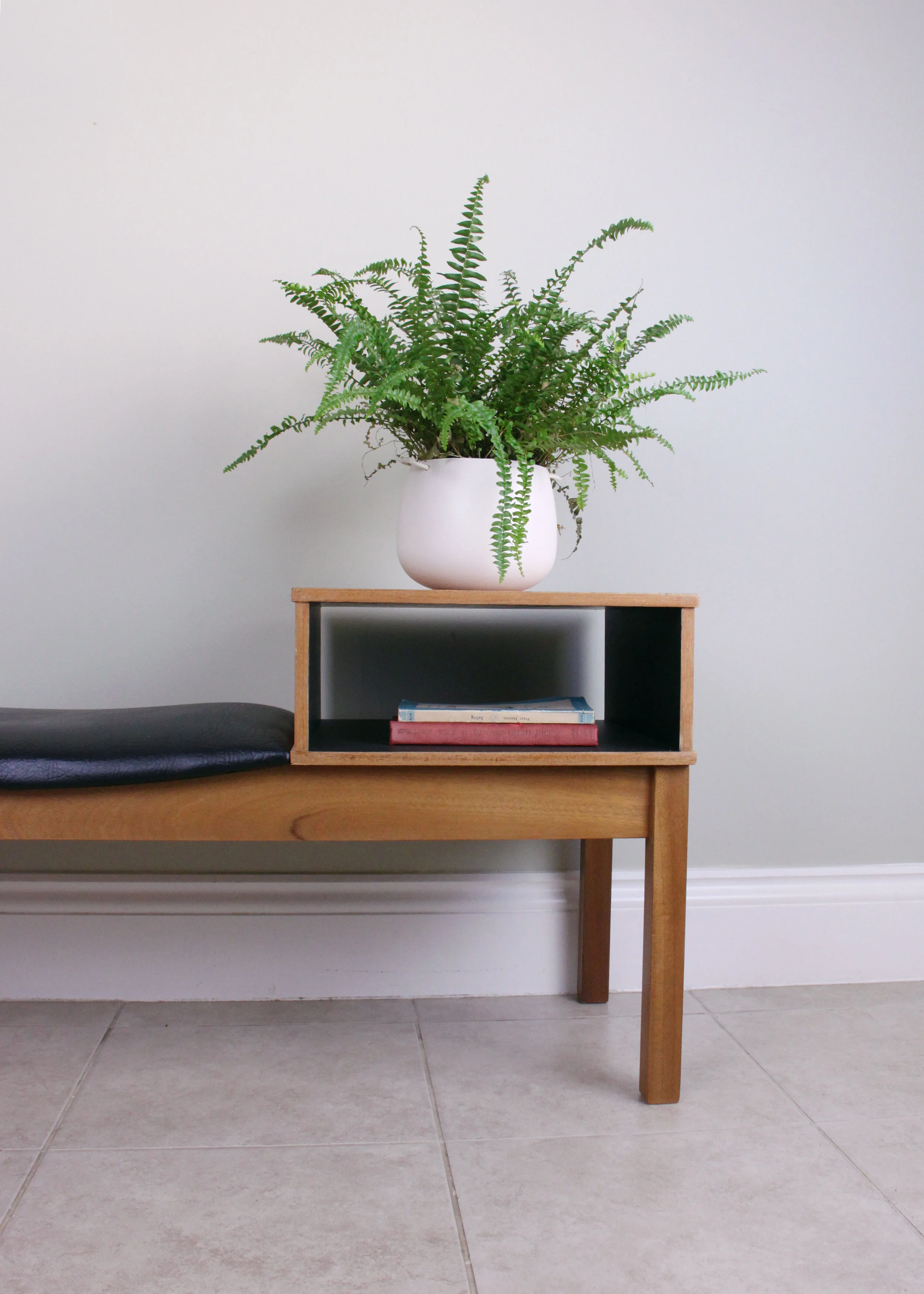 A potted fern on a wooden bench with a small shelf holding three books, against a plain white wall and tiled floor.