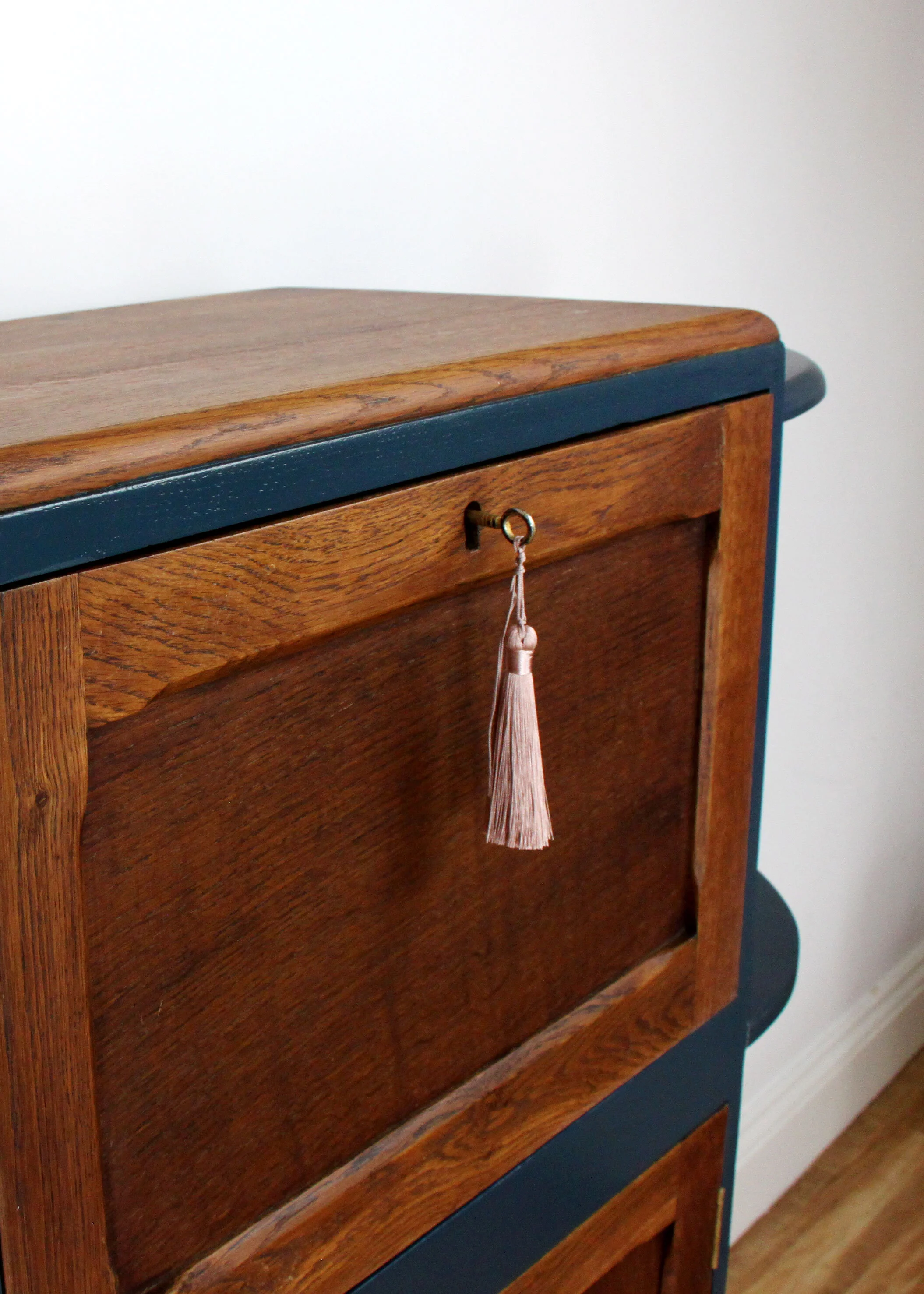 Close-up of a wooden cabinet with a dark blue trim, a brass keyhole with a pink tassel attached, and a plain white wall in the background.