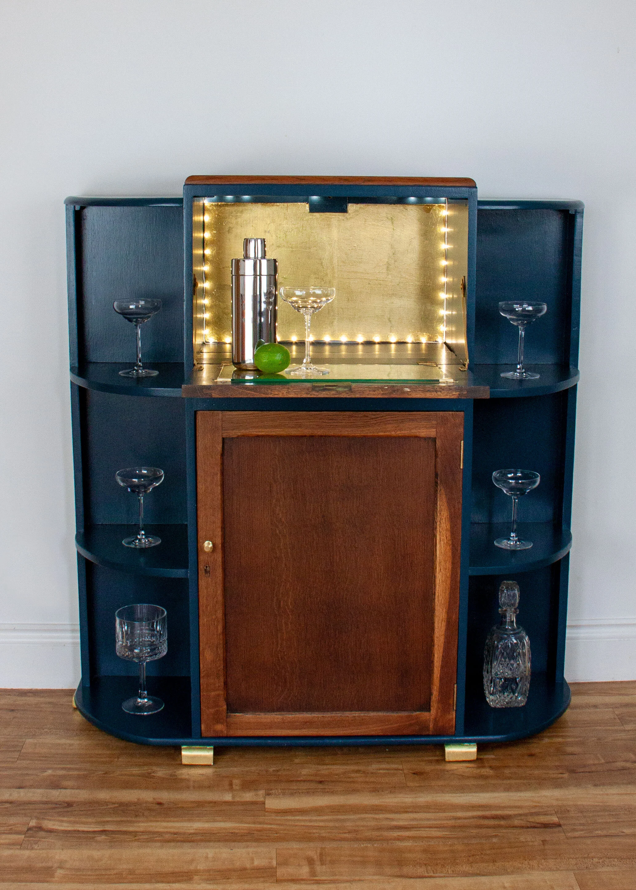 A vintage bar cabinet with a dark blue exterior, wooden sliding door, and built-in gold-lit display shelf holding cocktail shaker, glasses, a lime, and a decanter.