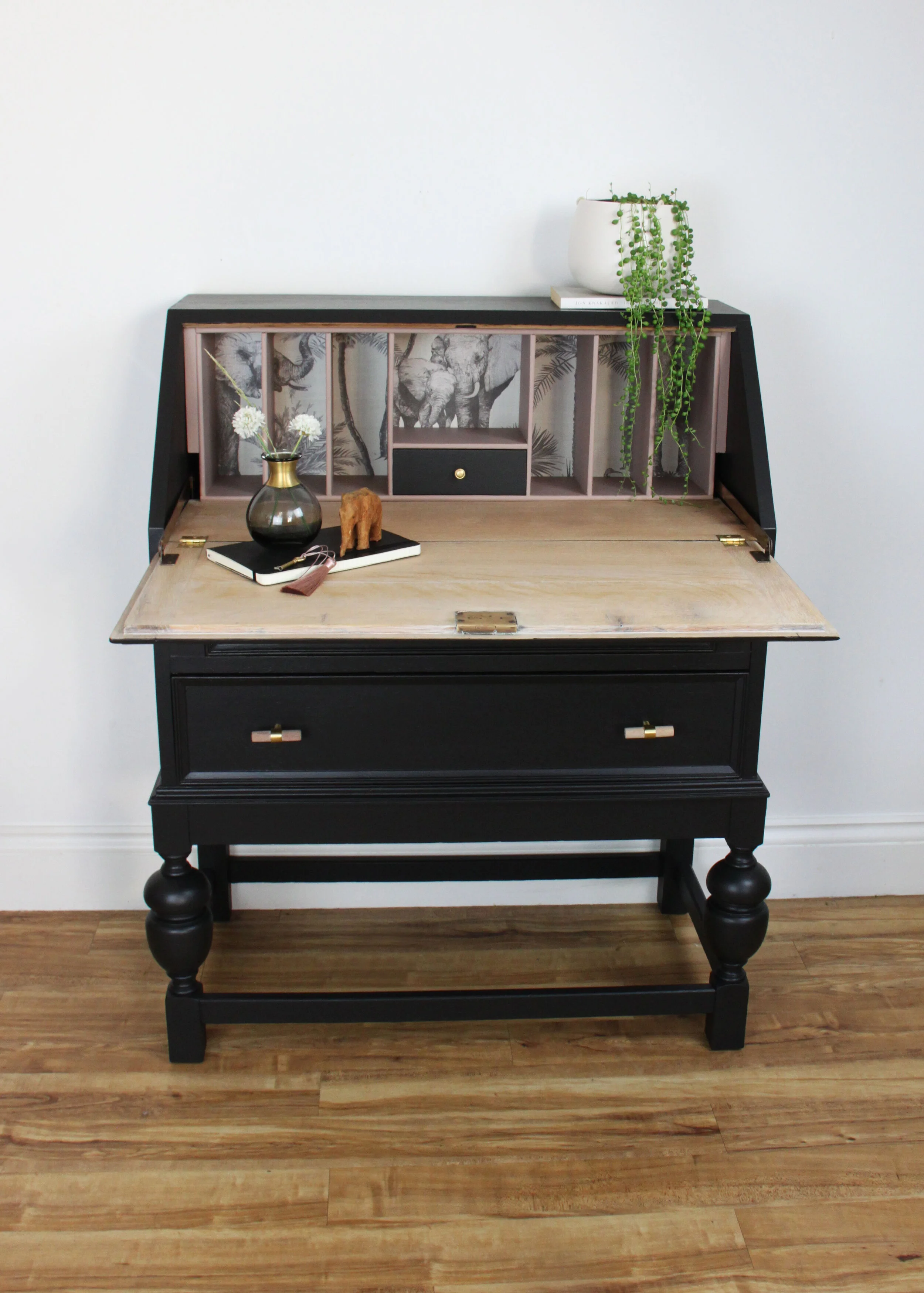 A black vintage writing desk with a fold-down writing surface, decorated with a black and white artwork background, a small drawer, and surrounded by a potted plant, a vase with flowers, and small decorative objects.
